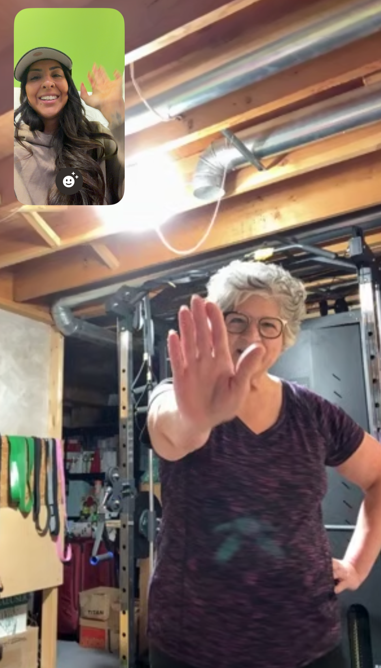 A woman with gray curly hair and glasses waving at the camera inside a garage or workshop, with shelves, tools, and equipment in the background; an inset video of a smiling woman in a cap and beige hoodie waving.