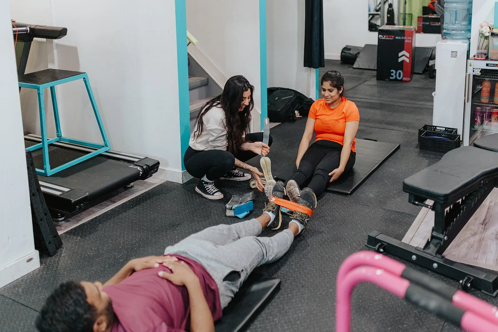 A woman assists a woman with a leg injury bandaged with orange strap while another person lies on a workout mat in a gym.