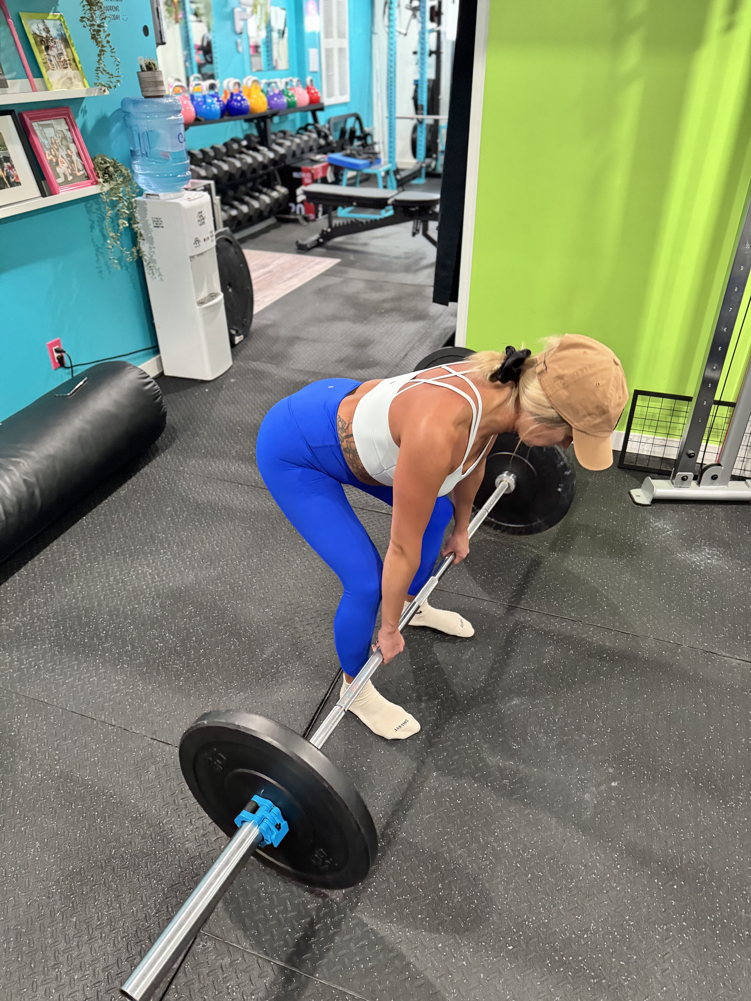 A woman in workout clothes lifting a barbell in a gym. The gym has colorful kettlebells, weights, and mirrors on the wall.
