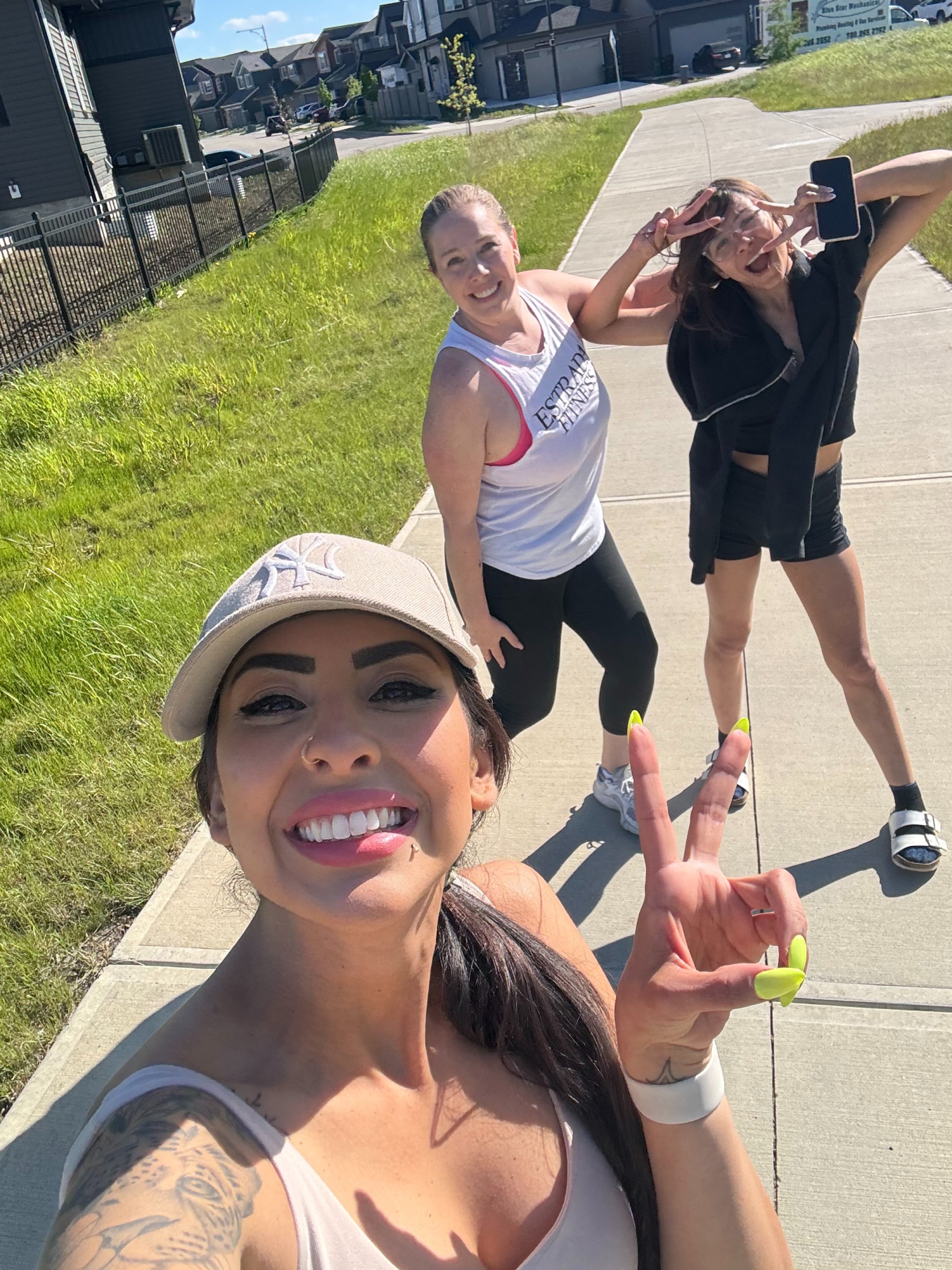 Three women taking a selfie outdoors on a sunny day, with one woman in the foreground and two women in the background making peace signs. They are on a sidewalk with houses and grass in the background.