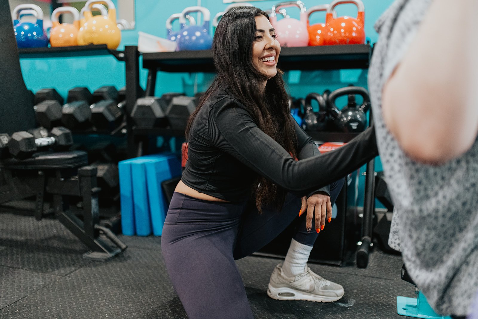A woman with long dark hair and a black athletic top is smiling while placing her hand on a person's shoulder inside a gym. The gym has colorful kettlebells, dumbbells, and other workout equipment on black racks in the background.