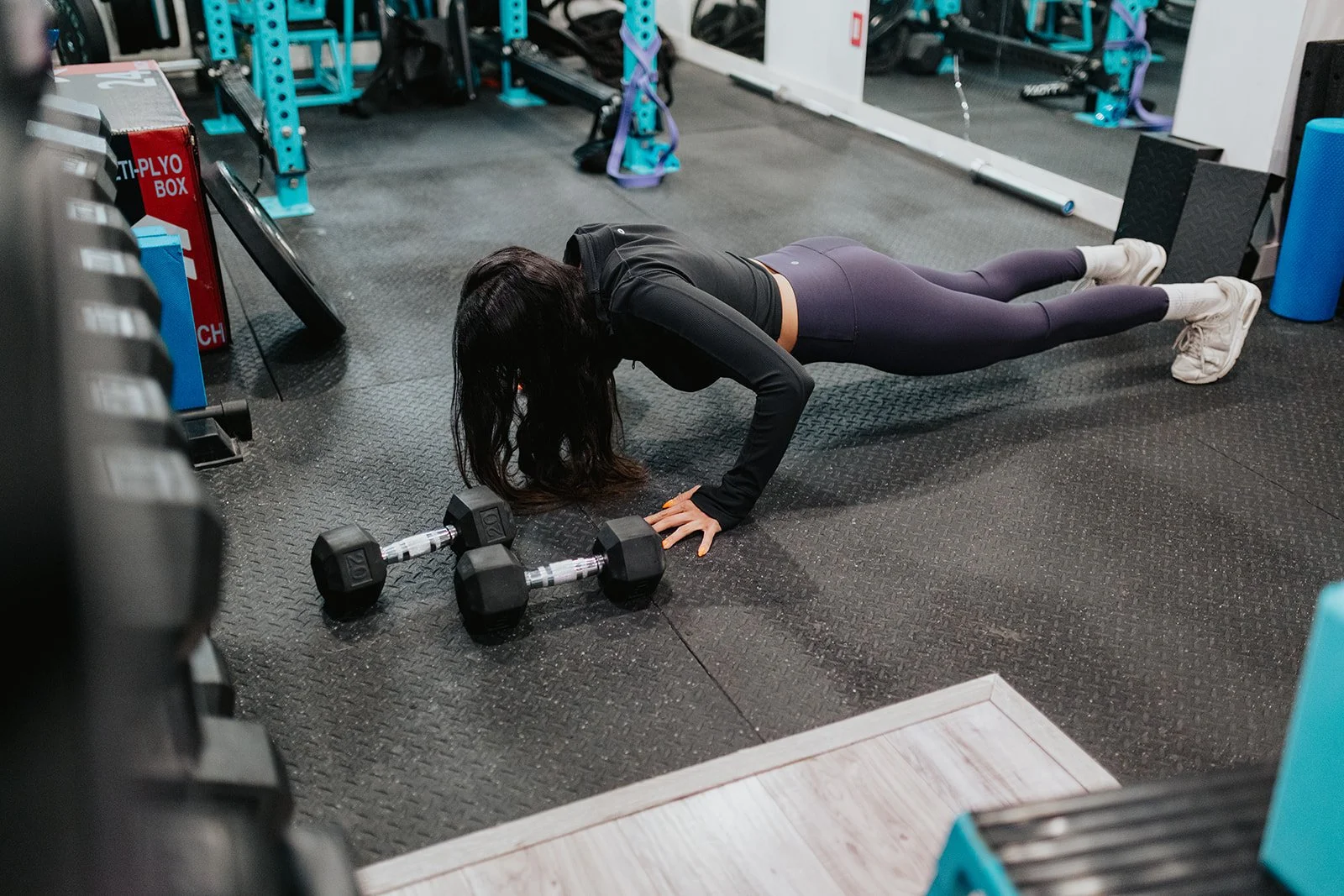 A woman performing push-ups on the gym floor next to a pair of dumbbells at a fitness center.
