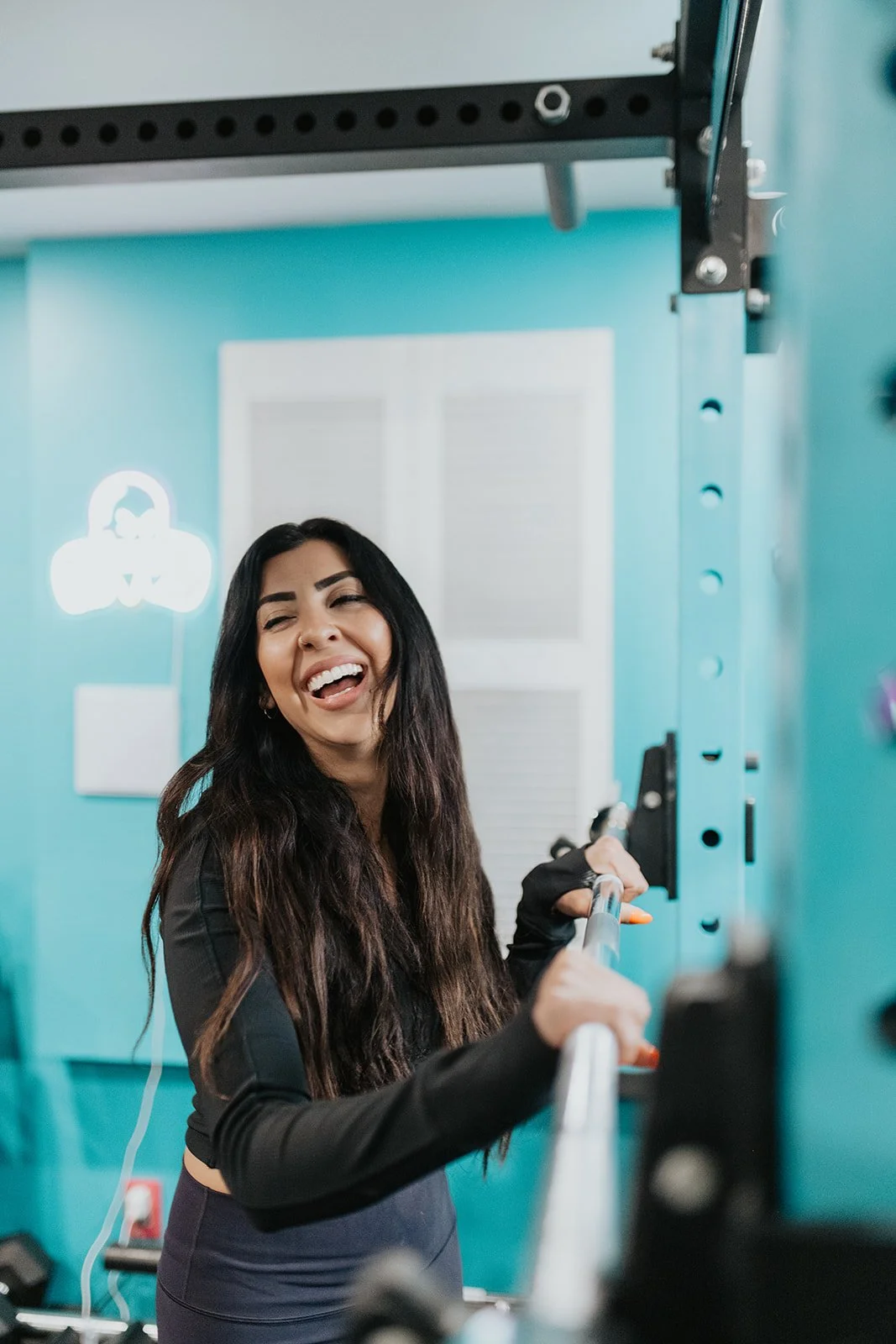 A woman with long dark hair smiling and holding onto a bar in a gym with light blue walls.