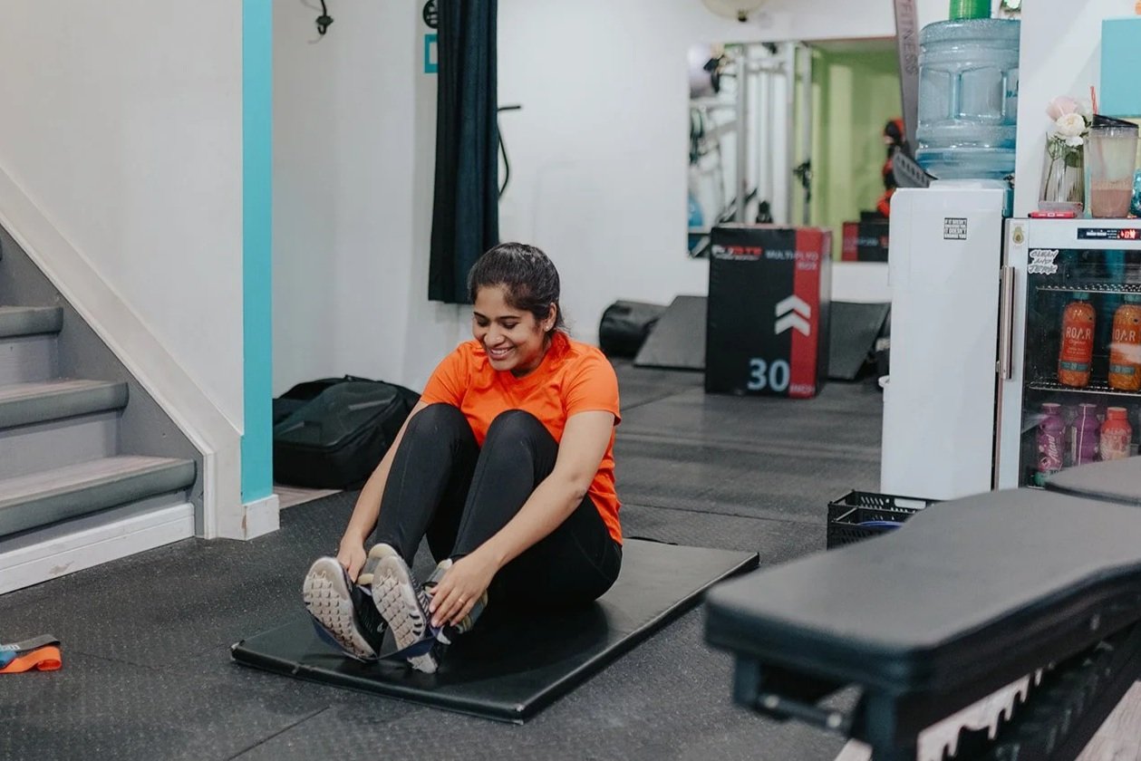 A young woman in an orange shirt and black pants sitting on a mat, tying her shoes in a gym or fitness room.