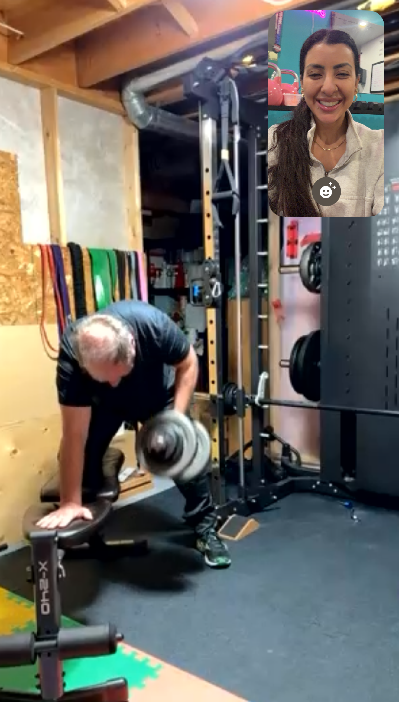 A man lifting a dumbbell with assistance from a woman in a gym setting. The gym has wooden beams, colorful resistance bands on the wall, and weightlifting equipment.
