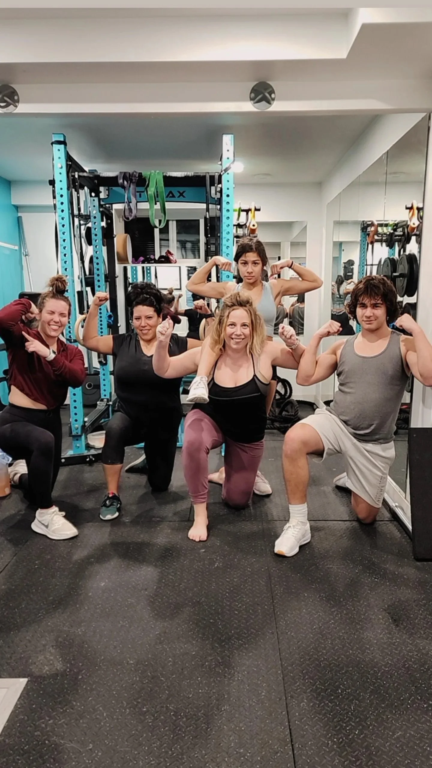 A group of five people, four women and one man, posing with flexed biceps in a gym, with exercise equipment visible in the background and a large mirror on the wall.