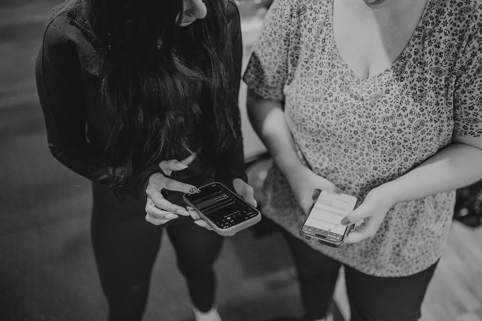 Two women standing close, looking at their smartphones, one with long curly hair and the other with a patterned top, in black and white.