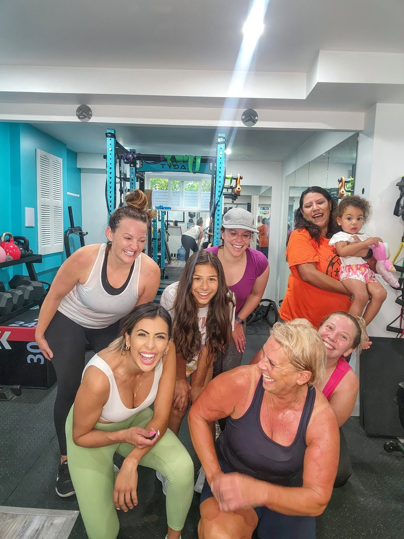 A group of women and a young girl smiling and posing in a gym. The women are wearing workout clothes, and one woman is holding a young girl. The gym has workout equipment and mirrors in the background.