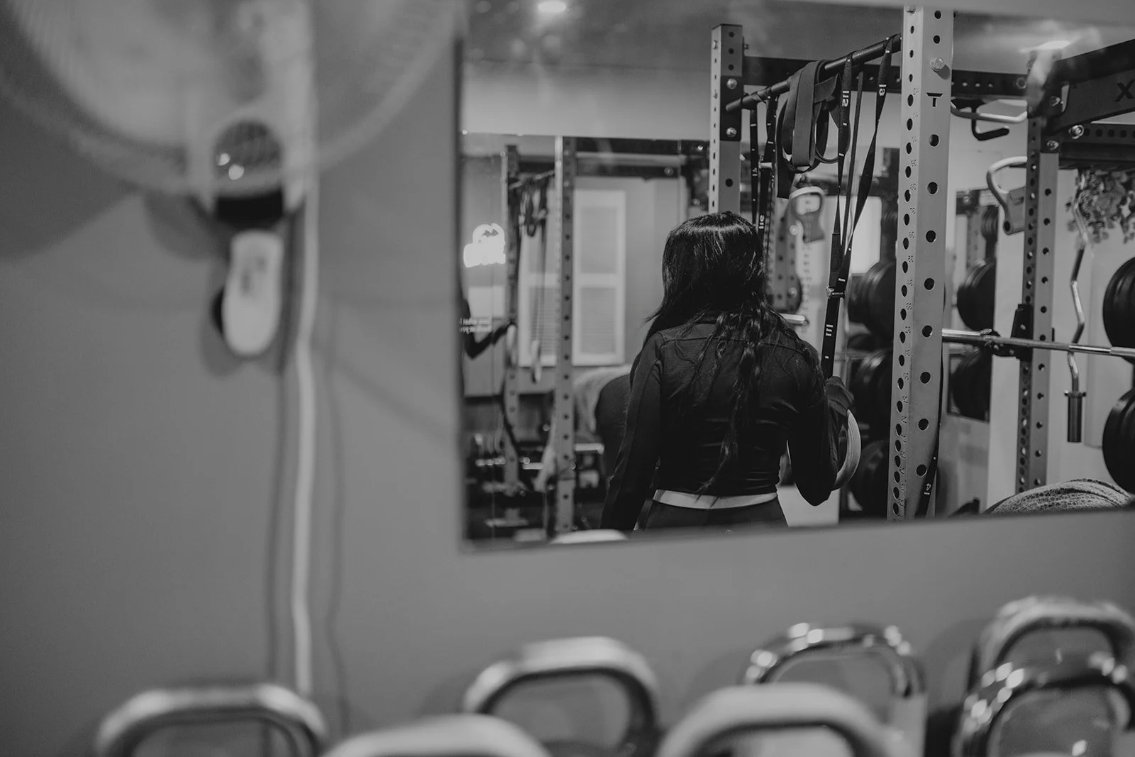 A woman with long dark hair in athletic clothing looks at weights in a gym, visible through a mirror.