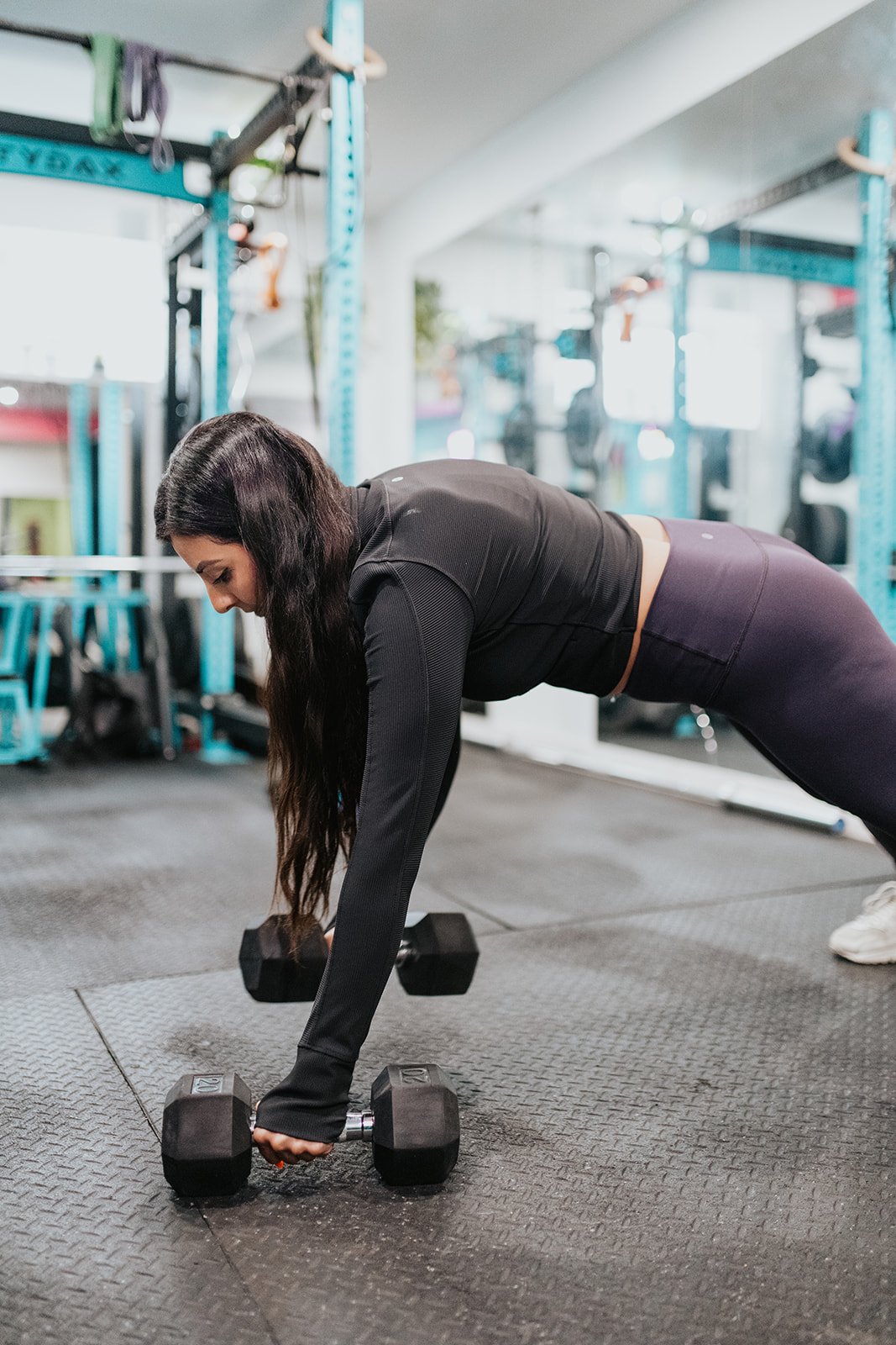 A woman performing a plank exercise with dumbbells in a gym, wearing a black long-sleeve top and dark leggings.