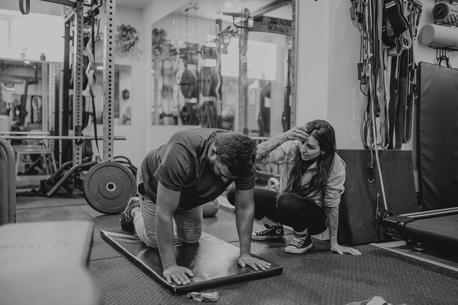 A woman assisting a man during a workout in a gym. The man is on a padded exercise mat, in a crawling position. The woman is kneeling beside him, holding his head and providing encouragement. The gym equipment can be seen in the background.