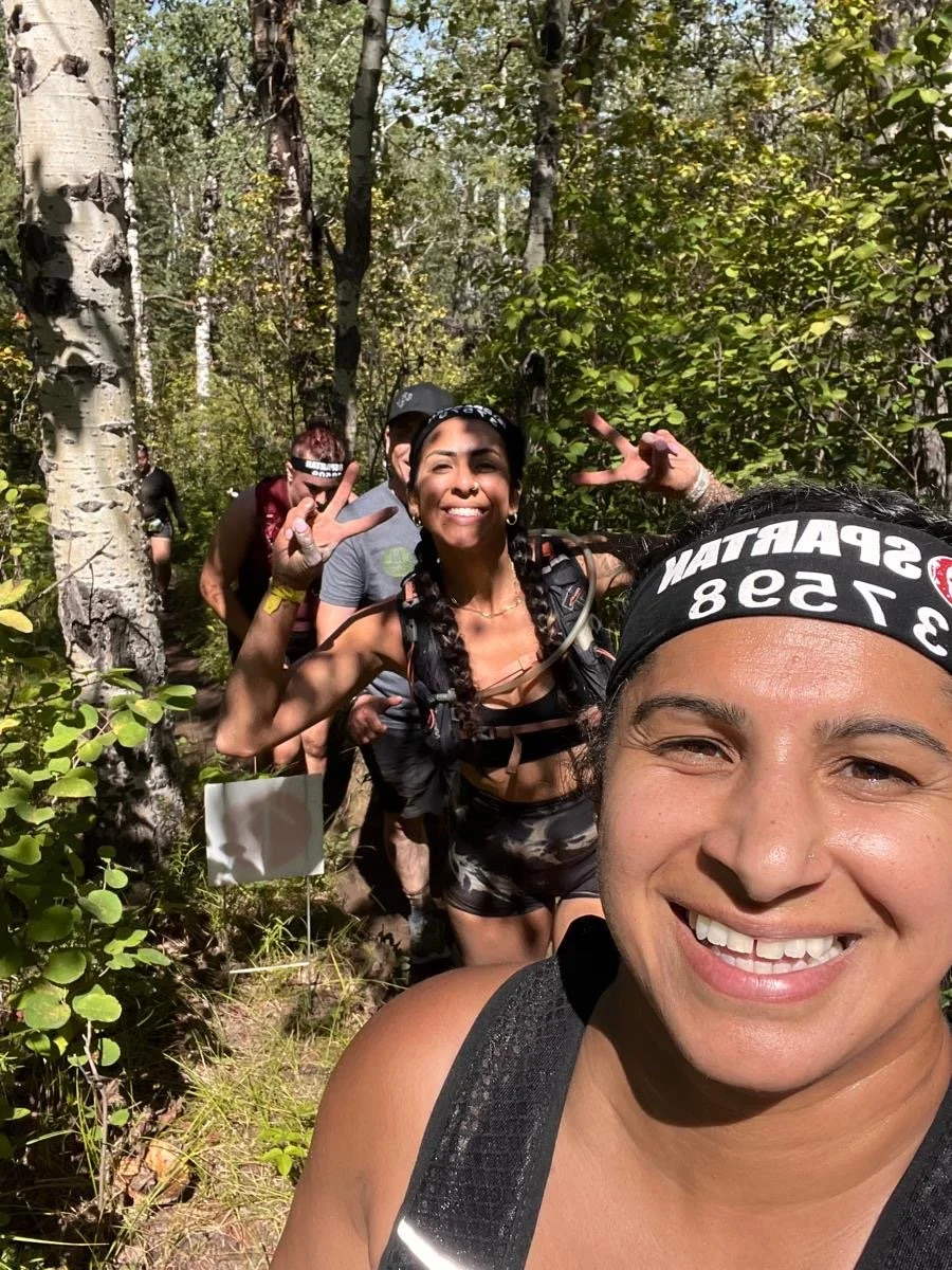 A group of people smiling and posing for a selfie while hiking through a lush forest. The woman in the foreground is taking the photo, with a headband that has the words 'SPARTAN 1538'. Others are making peace signs and smiling in the background.