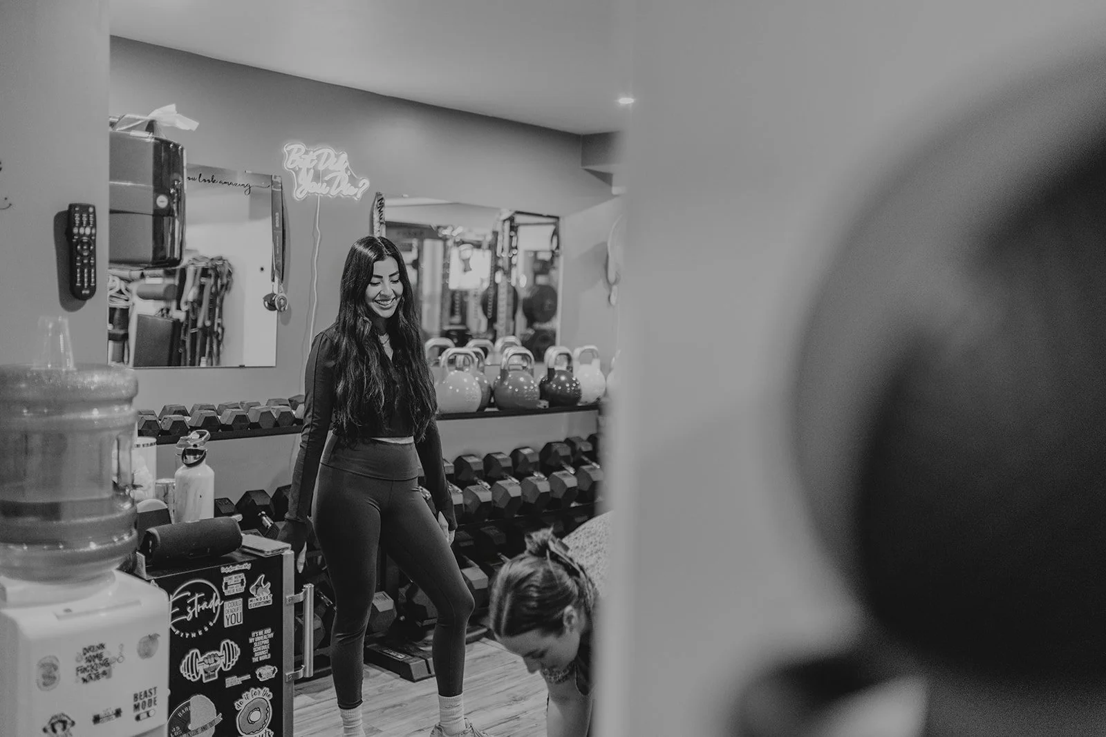 A woman with long dark hair smiling inside a gym, surrounded by kettlebells and dumbbells, with fitness equipment and a mirror in the background.