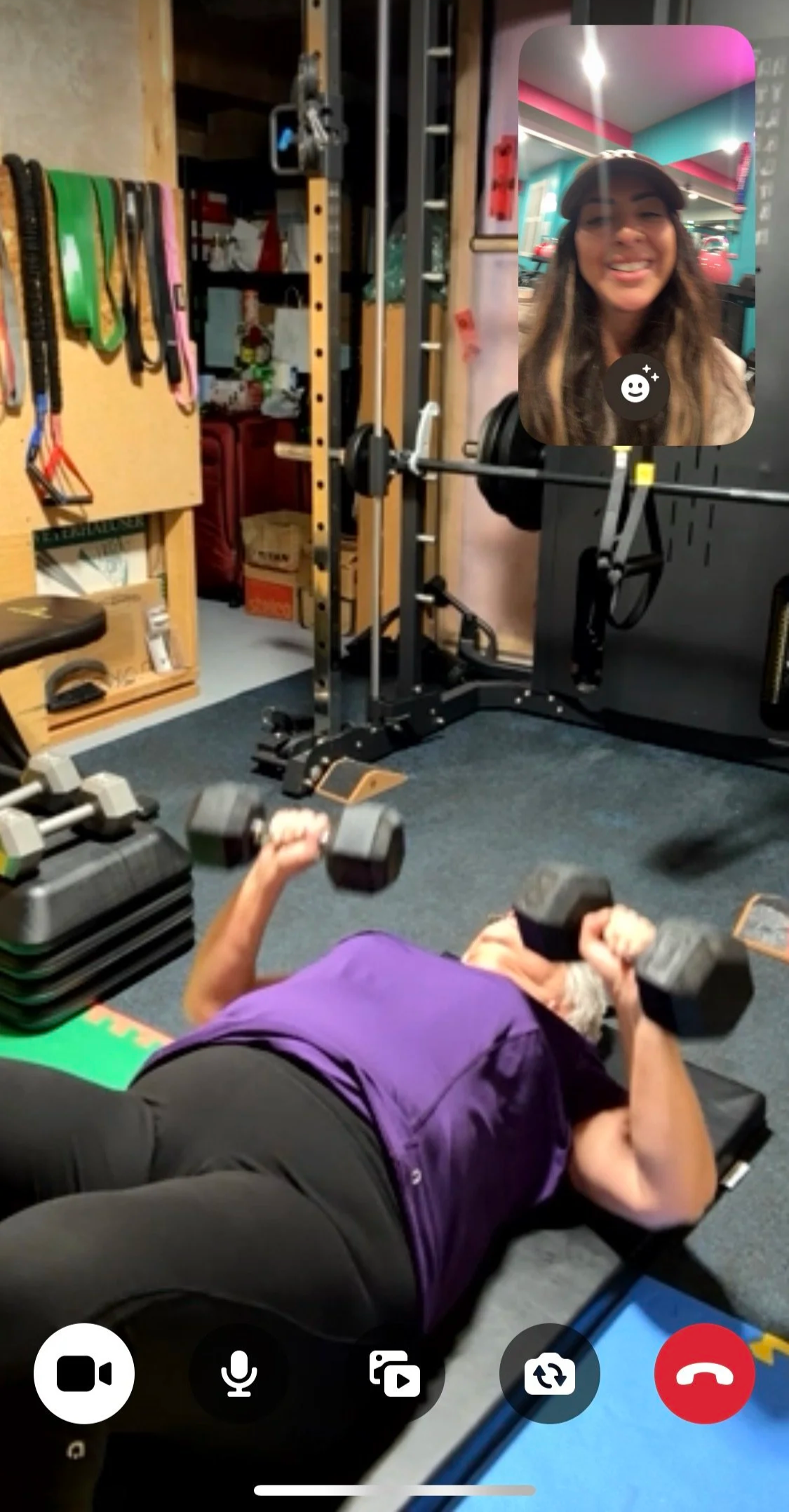 A woman lifting dumbbells while lying on a workout mat in a gym. A young woman is visible in a small screen overlay, smiling and wearing a cap.