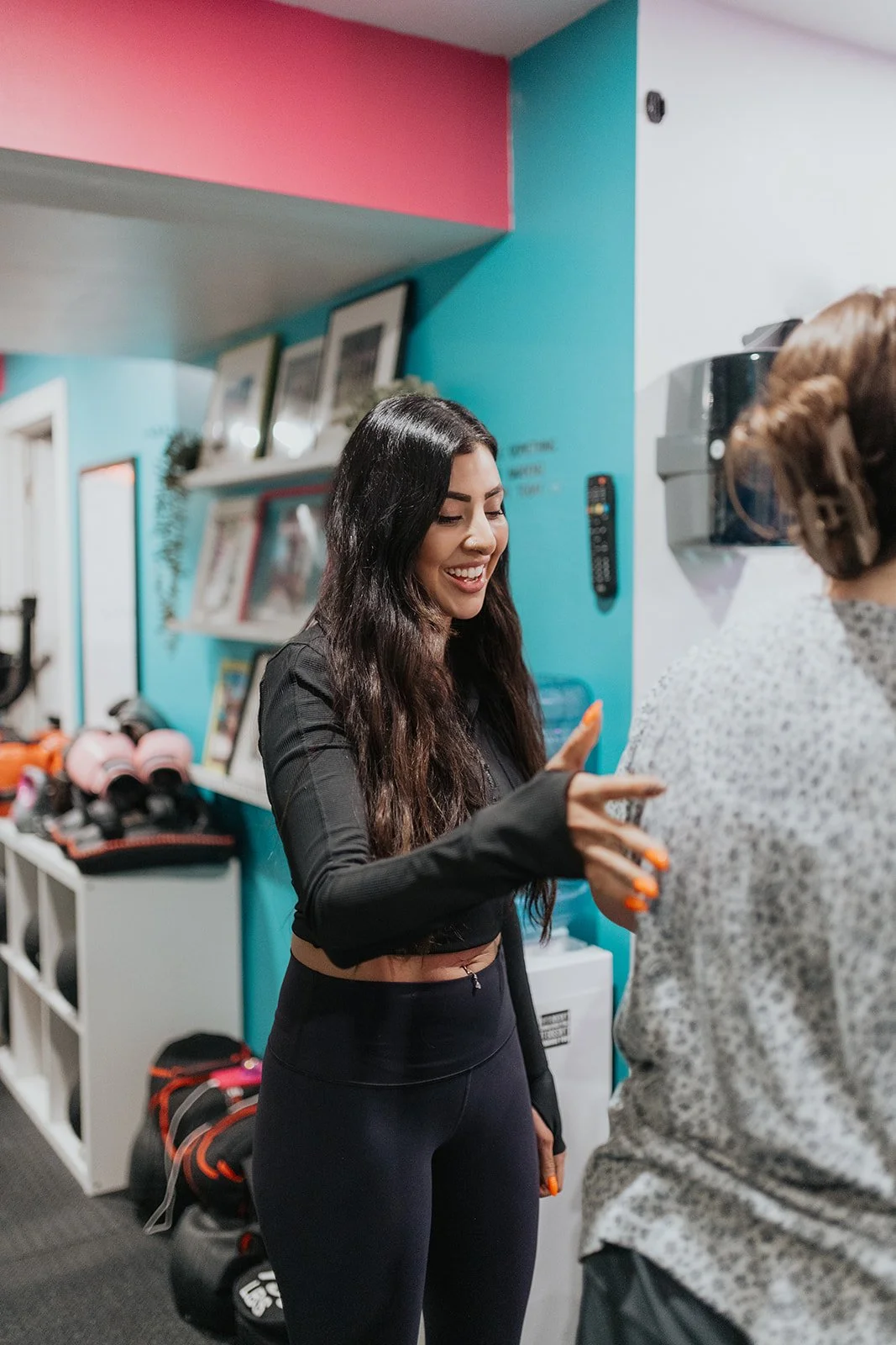 A woman with long dark hair smiling and talking to another person wearing headphones inside a colorful gym or fitness studio.