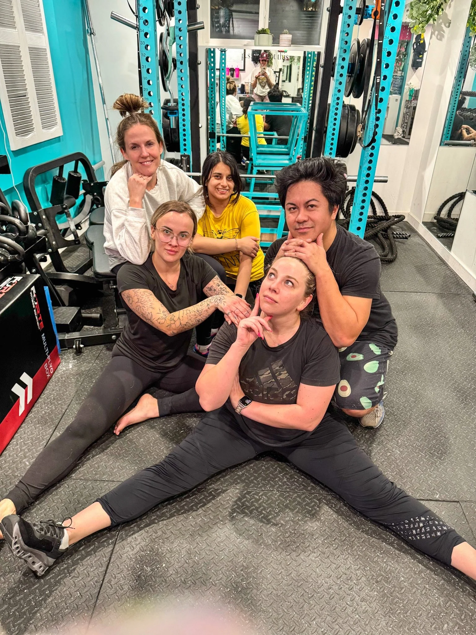 Group of five women in a gym, some sitting and some lying down, with gym equipment and mirrors in the background.