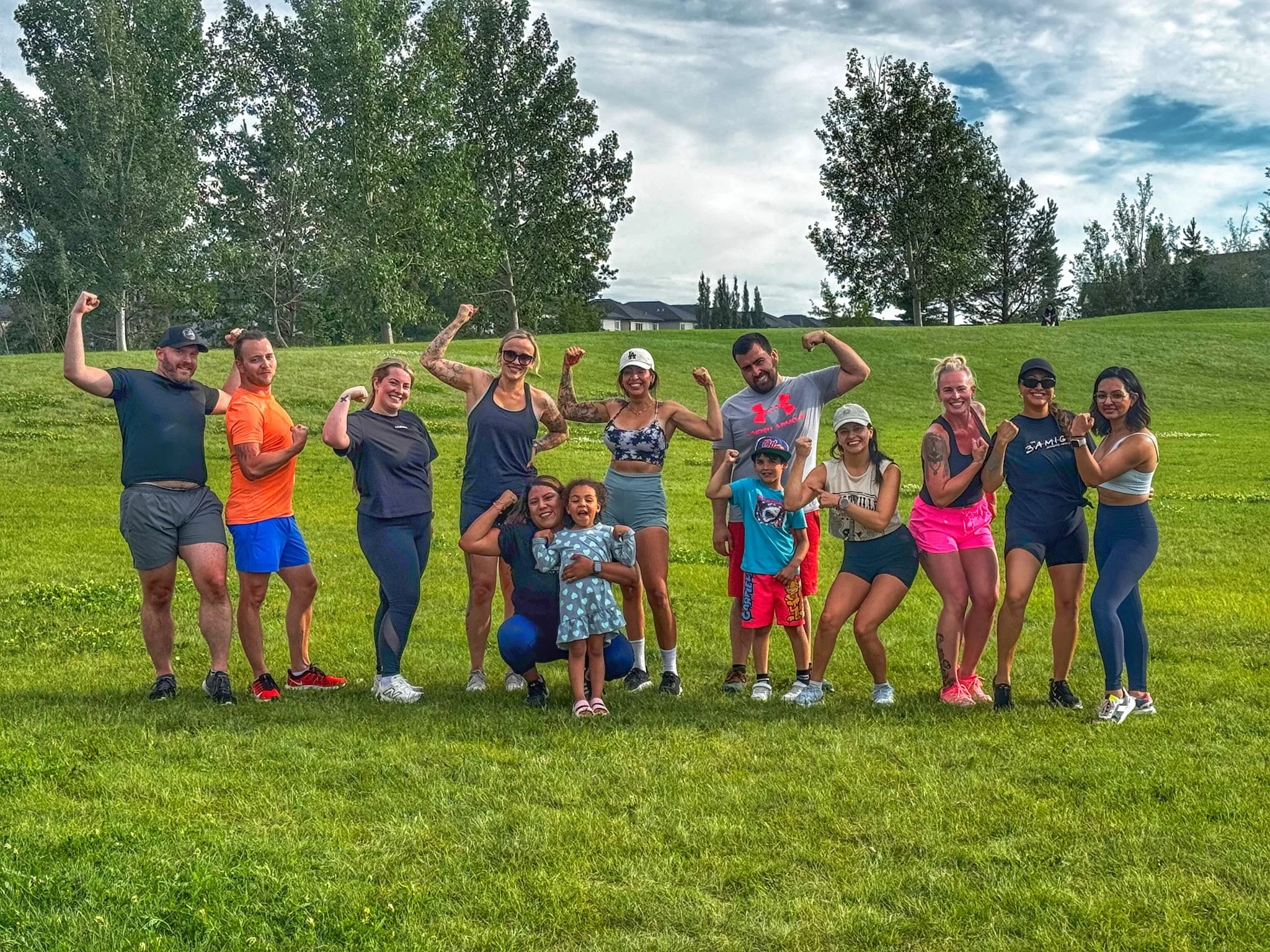 A diverse group of people, including adults and children, standing outdoors on grass, flexing their muscles and smiling, with trees and a cloudy sky in the background.