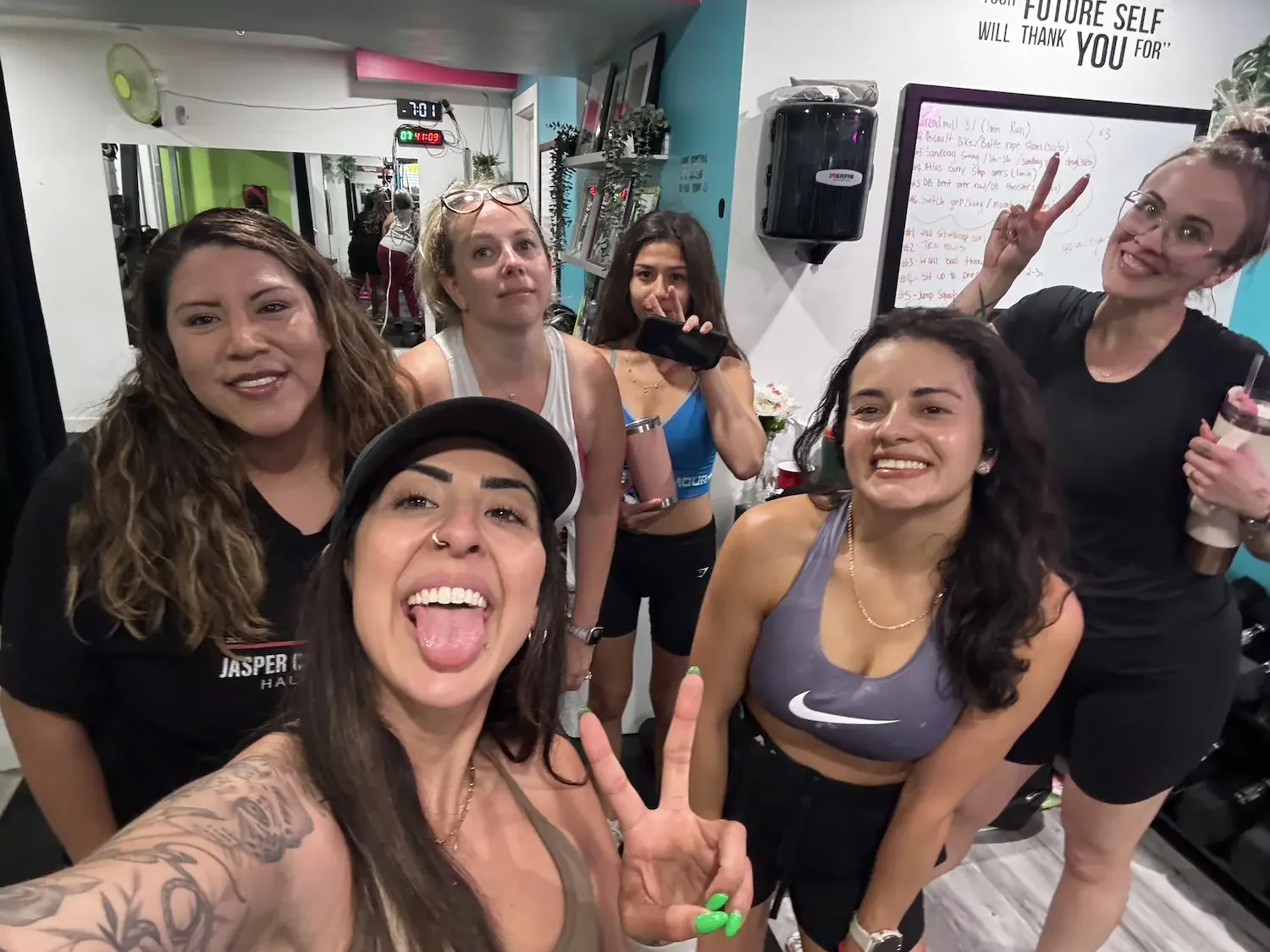 Group of women taking a selfie in a fitness studio. They are smiling and making peace signs. The studio has white walls, a whiteboard with writing, and a shelf with plants and framed pictures.