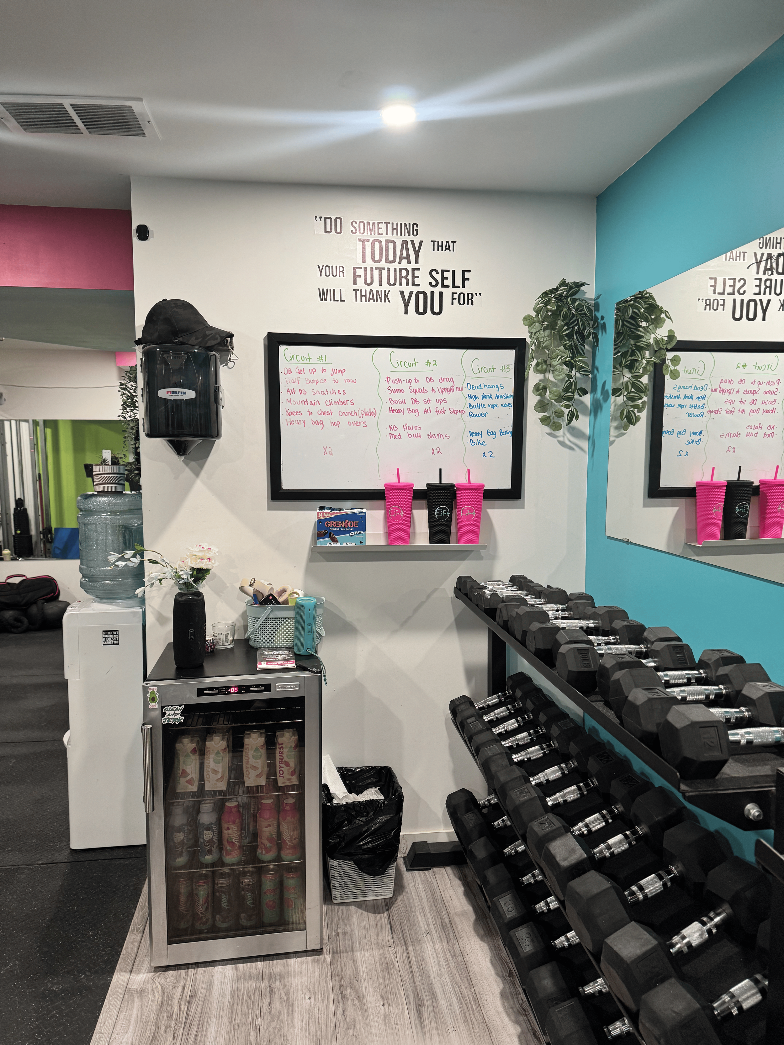 View of a gym corner with a white fridge holding beverages, a water dispenser on top, a mini refrigerator below, a trash can, a whiteboard with workout circuits, a rack of black and gray dumbbells, and pink and black water bottles on a wall-mounted shelf. There is a motivational quote on the wall and indoor plants.