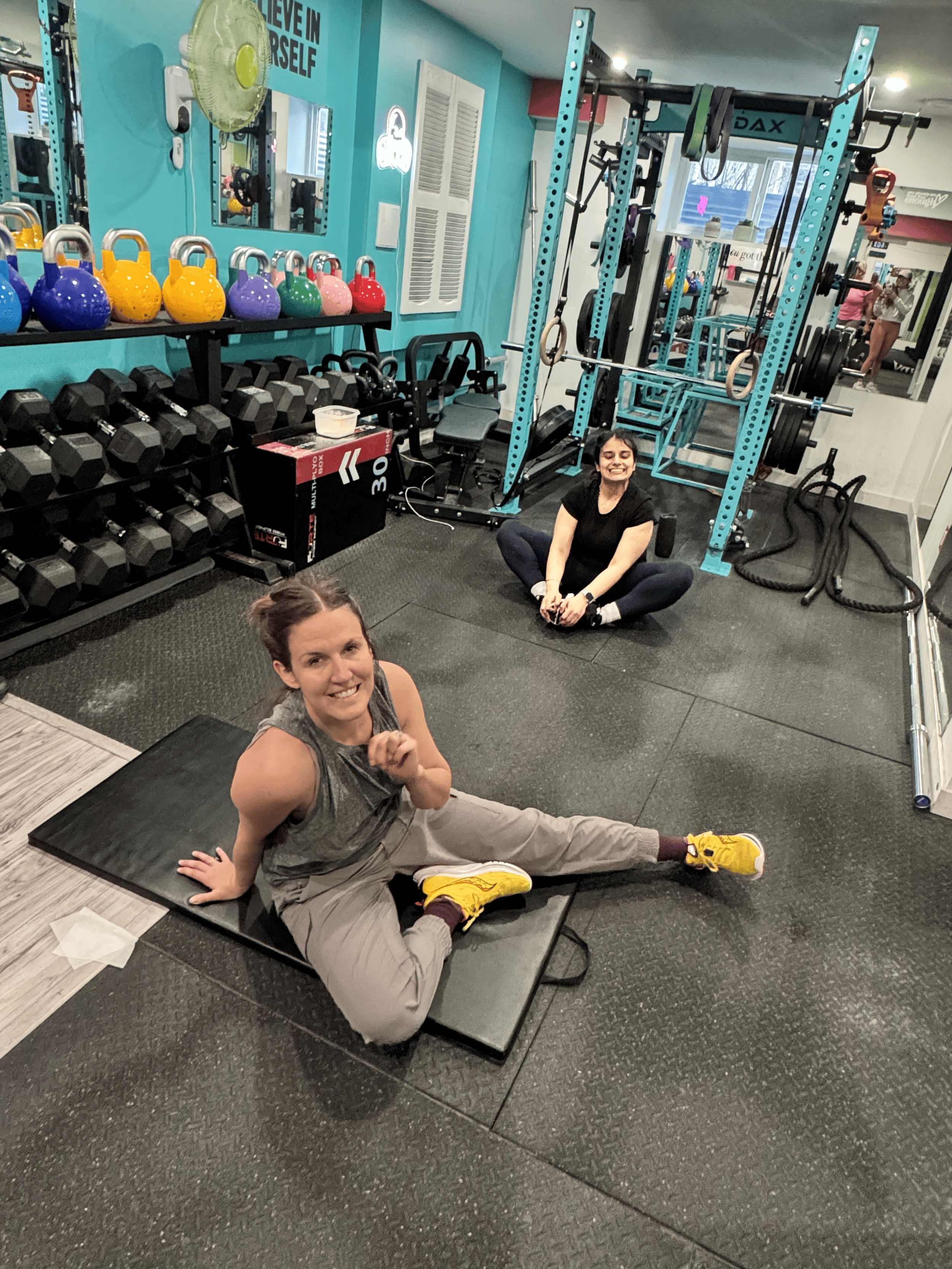 Two women, one sitting on the floor stretching and the other sitting behind her, smiling at the gym with weightlifting equipment and kettlebells in the background.