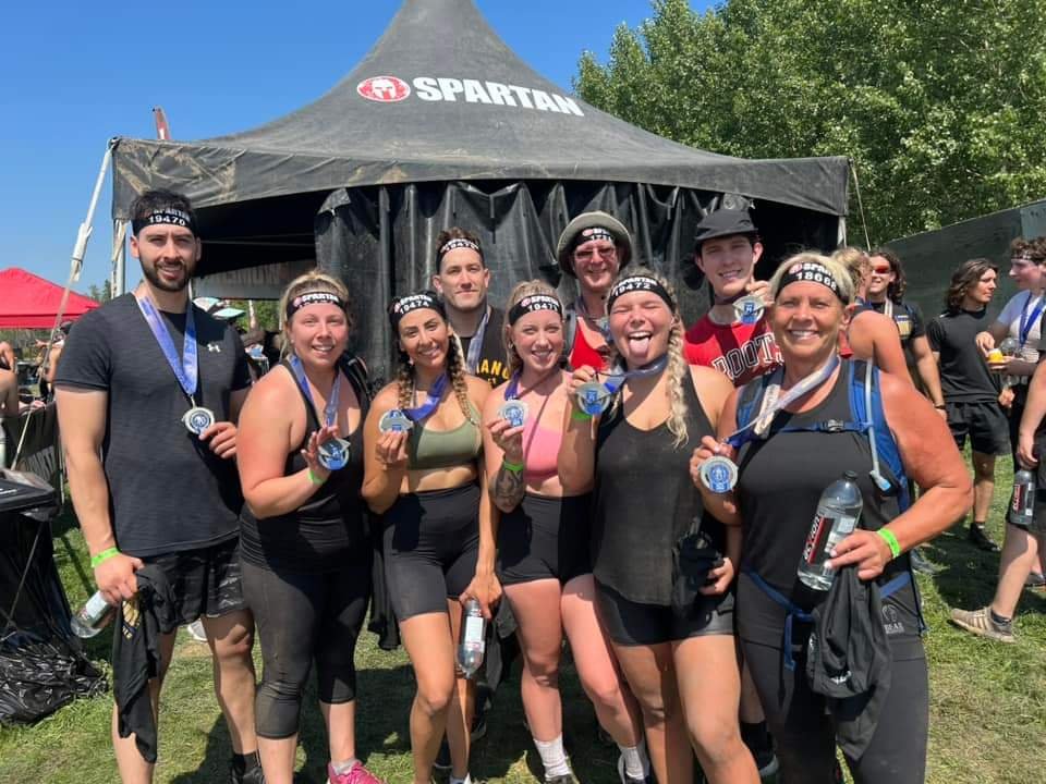 Group of people at Spartan obstacle course event, wearing medals and athletic gear, smiling with a tent in the background.