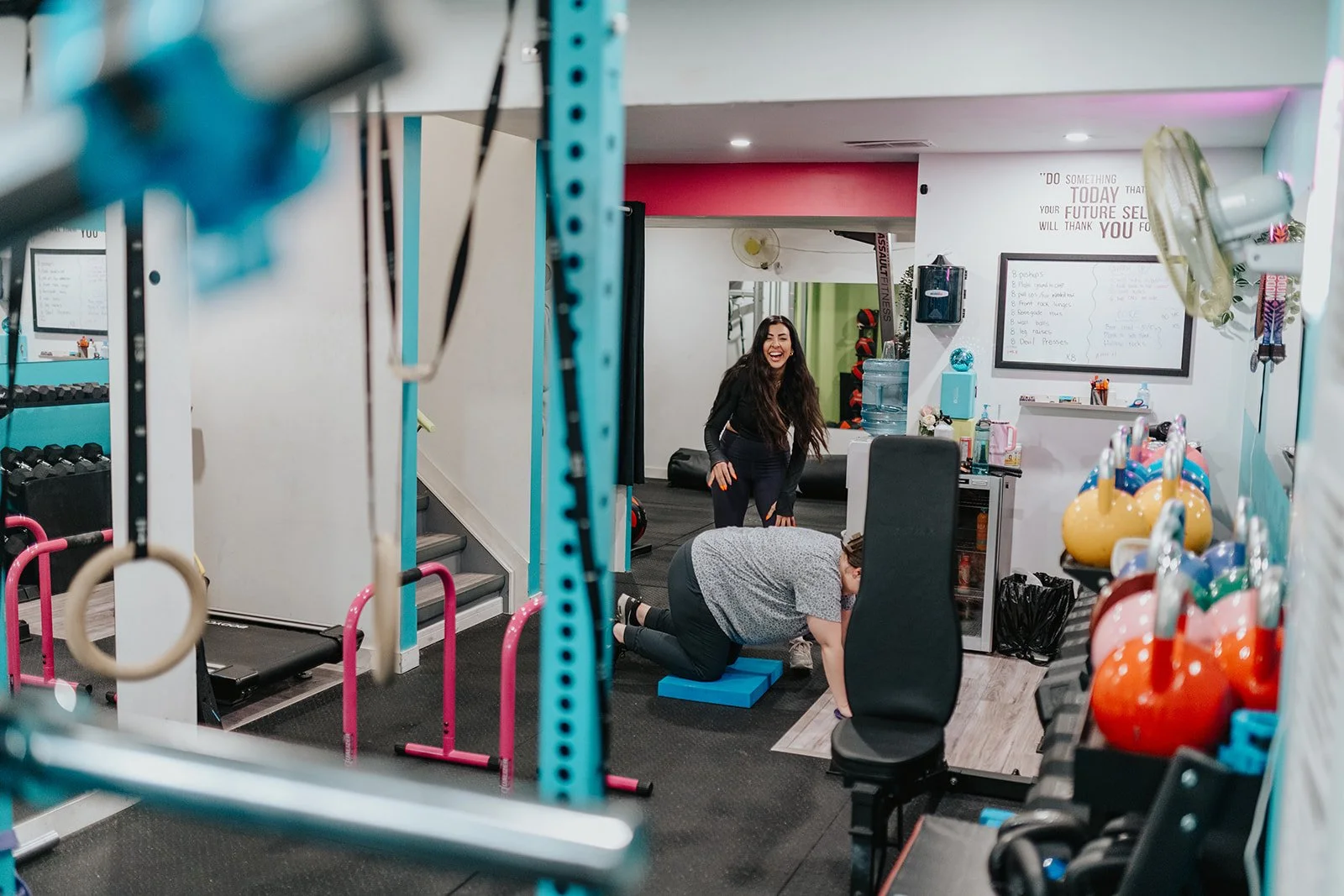 A fitness gym with two women working out, one is lying on a foam pad and the other is smiling nearby. The gym has colorful kettlebells, workout equipment, and a whiteboard with a workout plan.