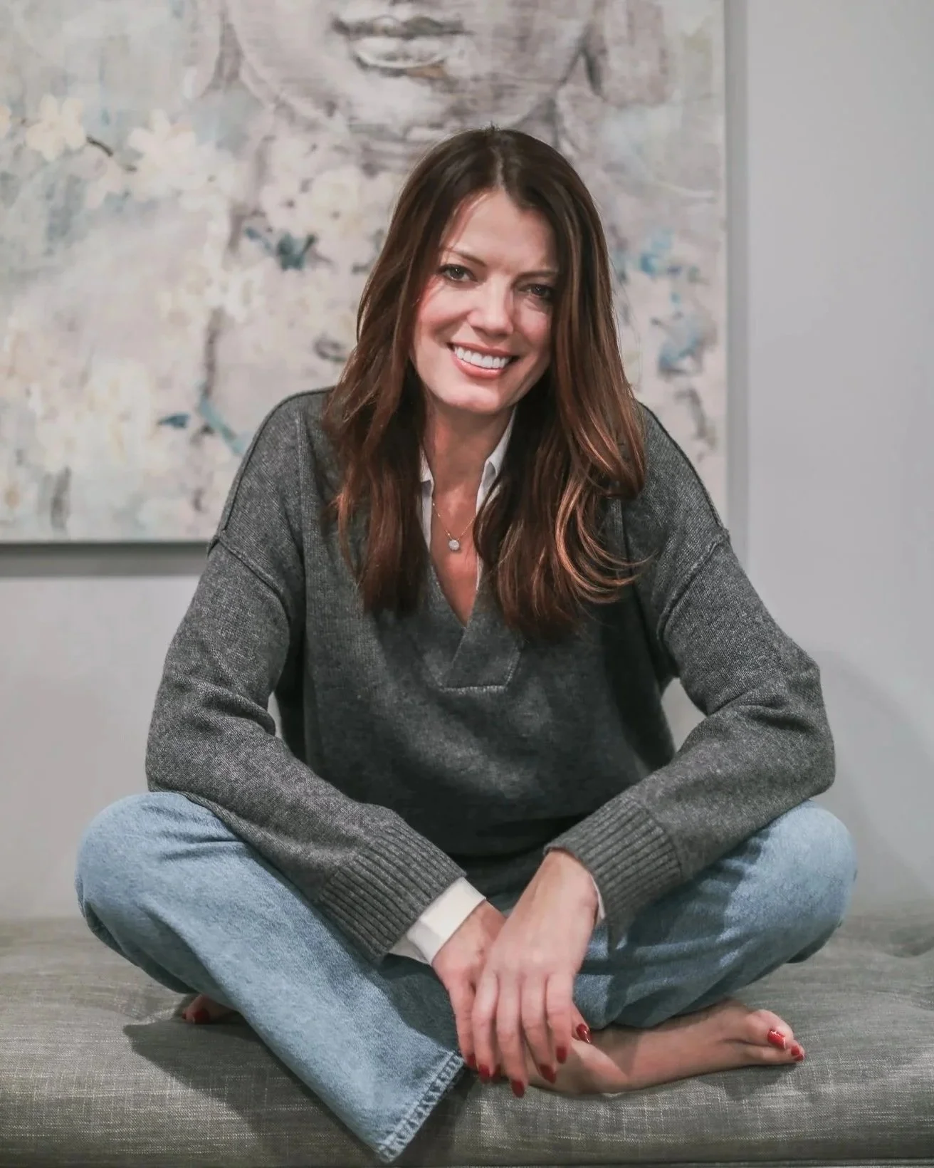 A woman with brown hair sitting cross-legged on a gray cushion in a room, smiling at the camera with a large abstract floral painting behind her.