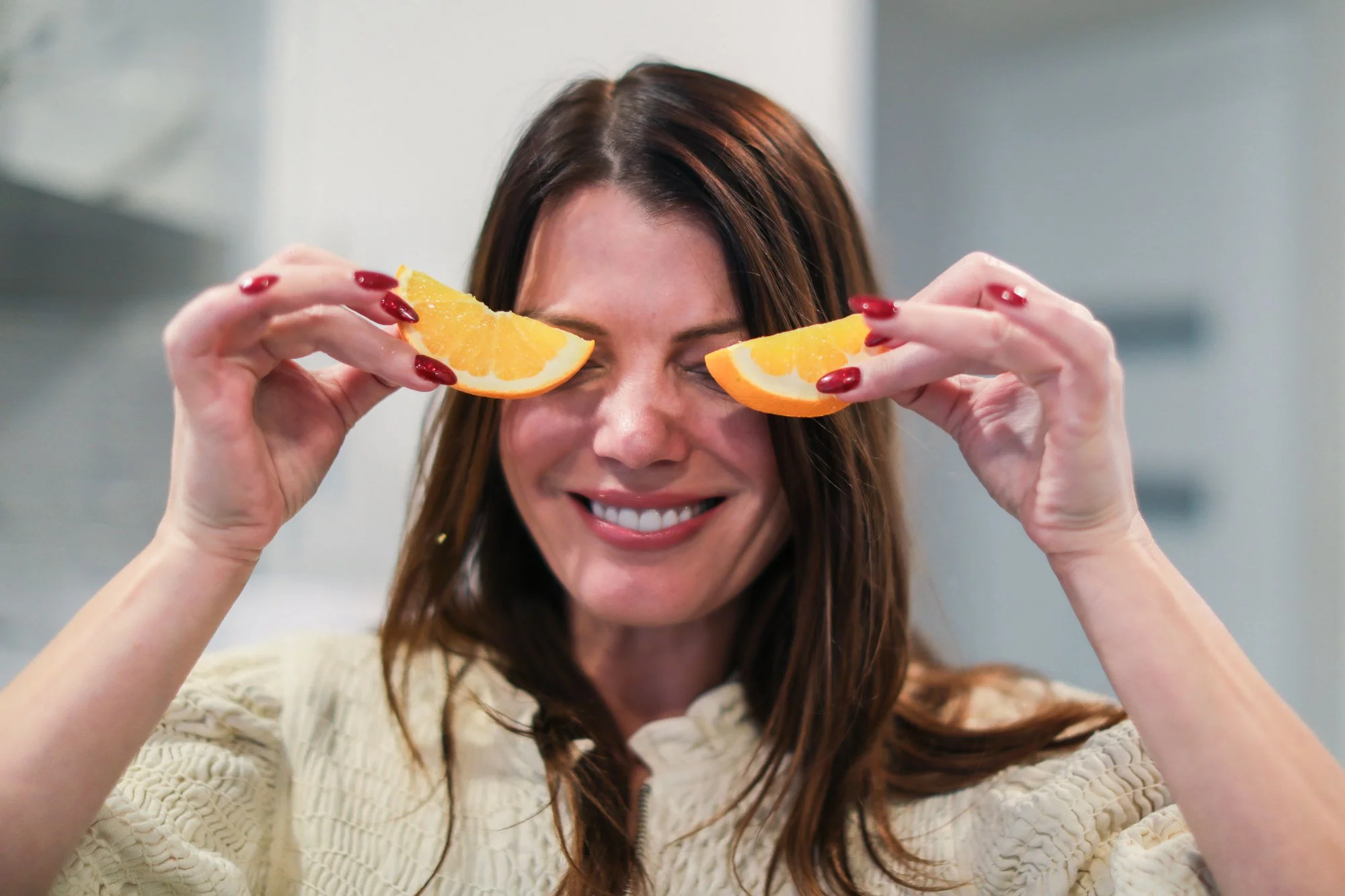 A woman with long brown hair and red nails smiling while holding orange slices over her eyes.