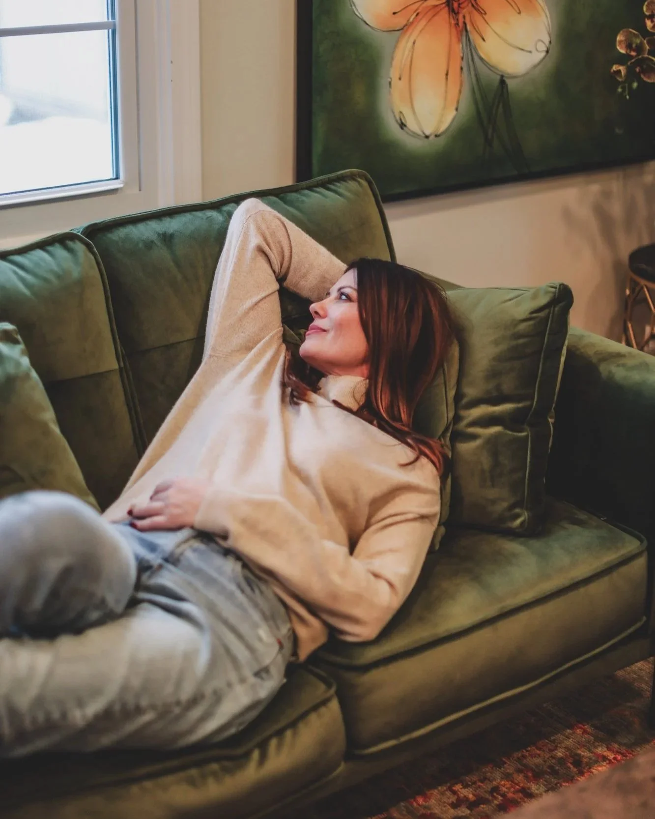 A woman with brown hair wearing a beige sweater resting on a green velvet couch with her arm behind her head, looking content.