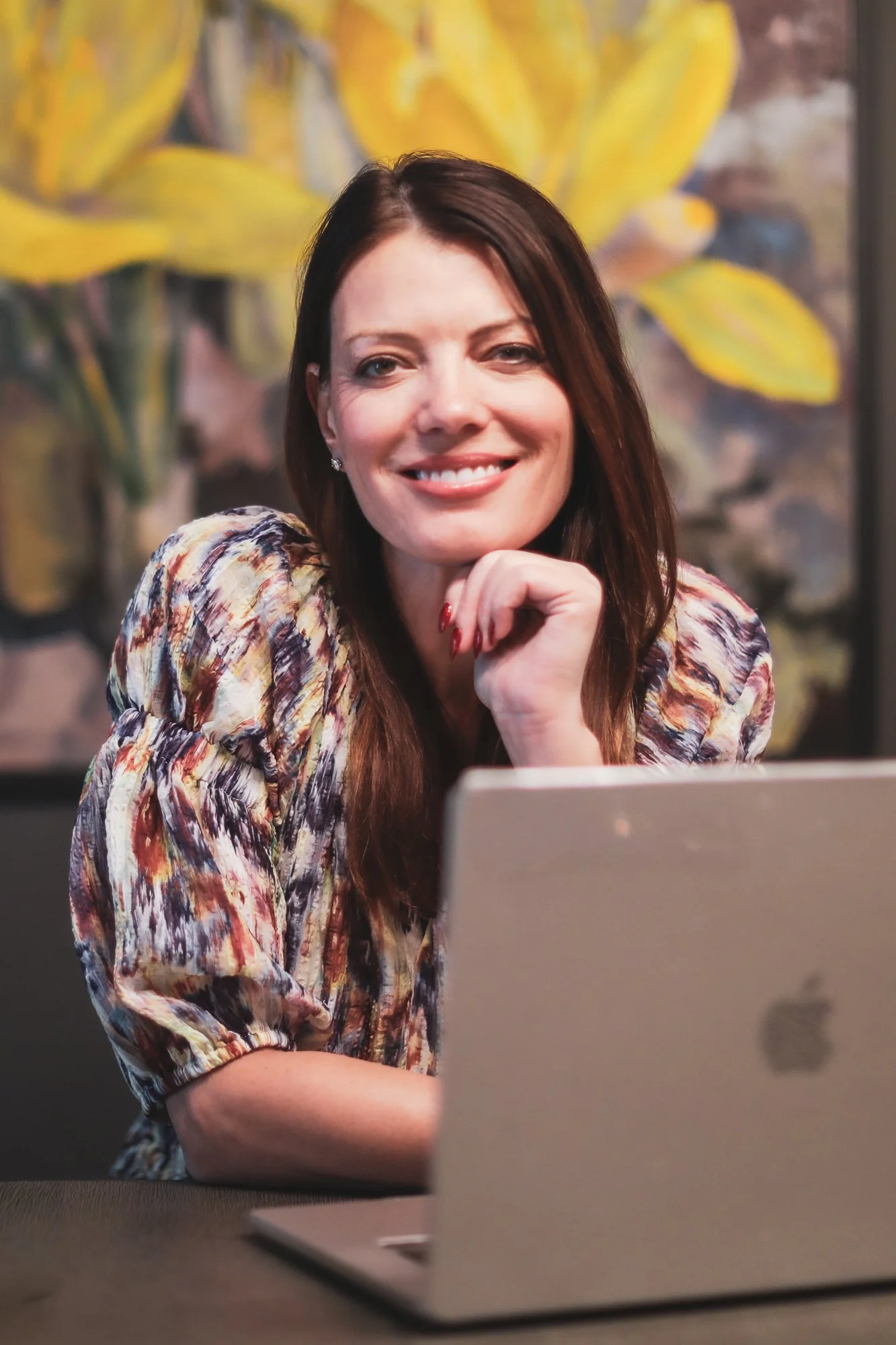 A woman with long brown hair, wearing a patterned blouse, smiling and resting her chin on her hand while sitting in front of a laptop with a blurred background of yellow and green leaves.