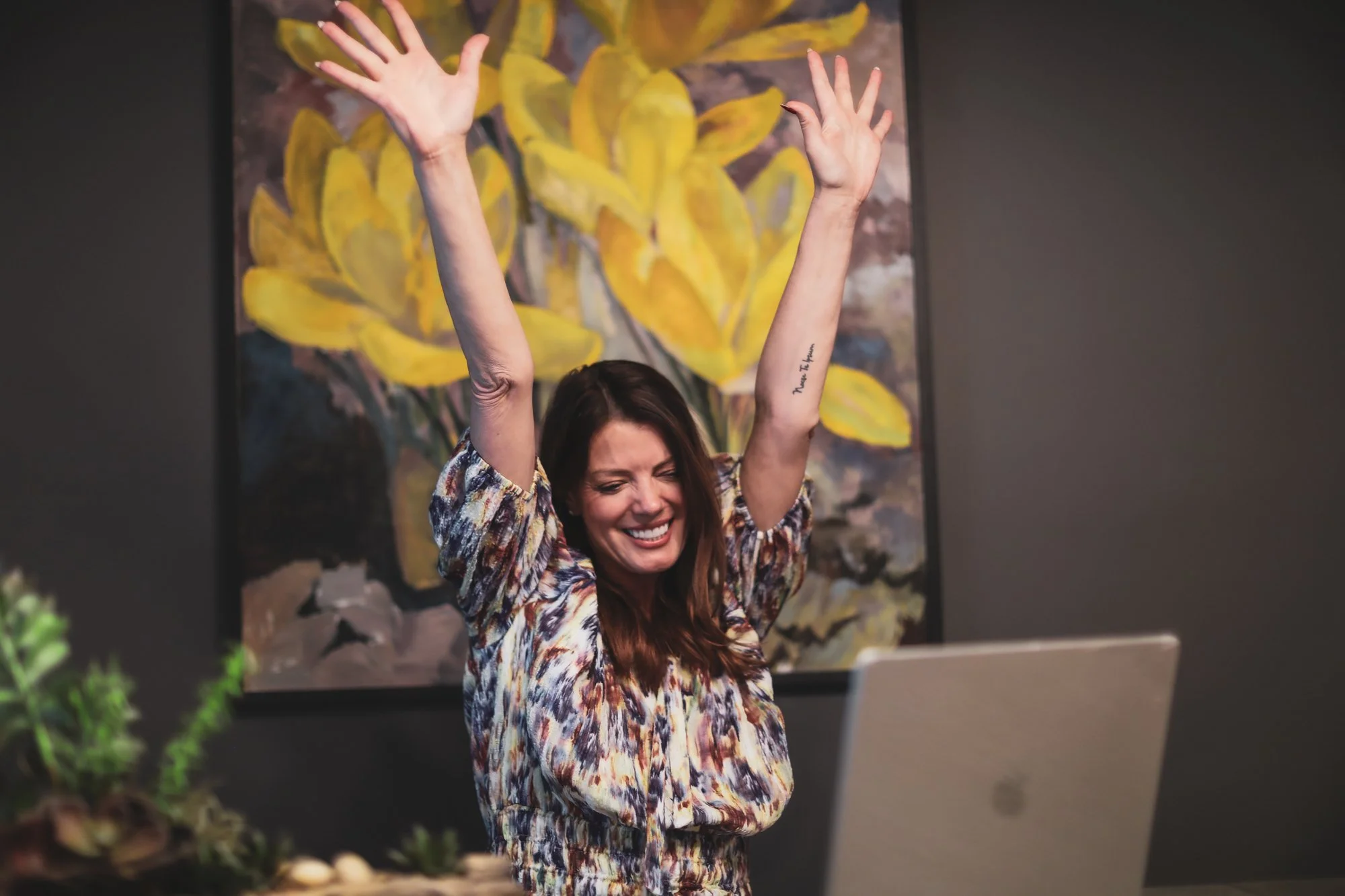 A woman with long brown hair is smiling with her eyes closed and arms raised, sitting in front of a laptop with a colorful abstract painting of yellow leaves behind her.