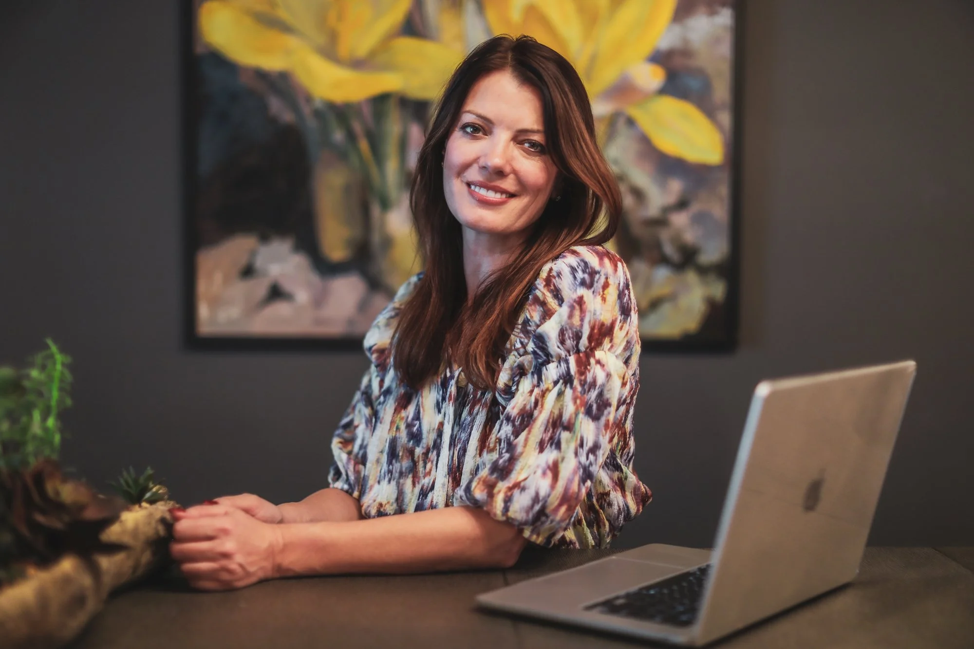 A woman with brown hair and a colorful patterned blouse sitting at a desk with a laptop, smiling at the camera. In the background, there is a yellow floral painting on the wall.