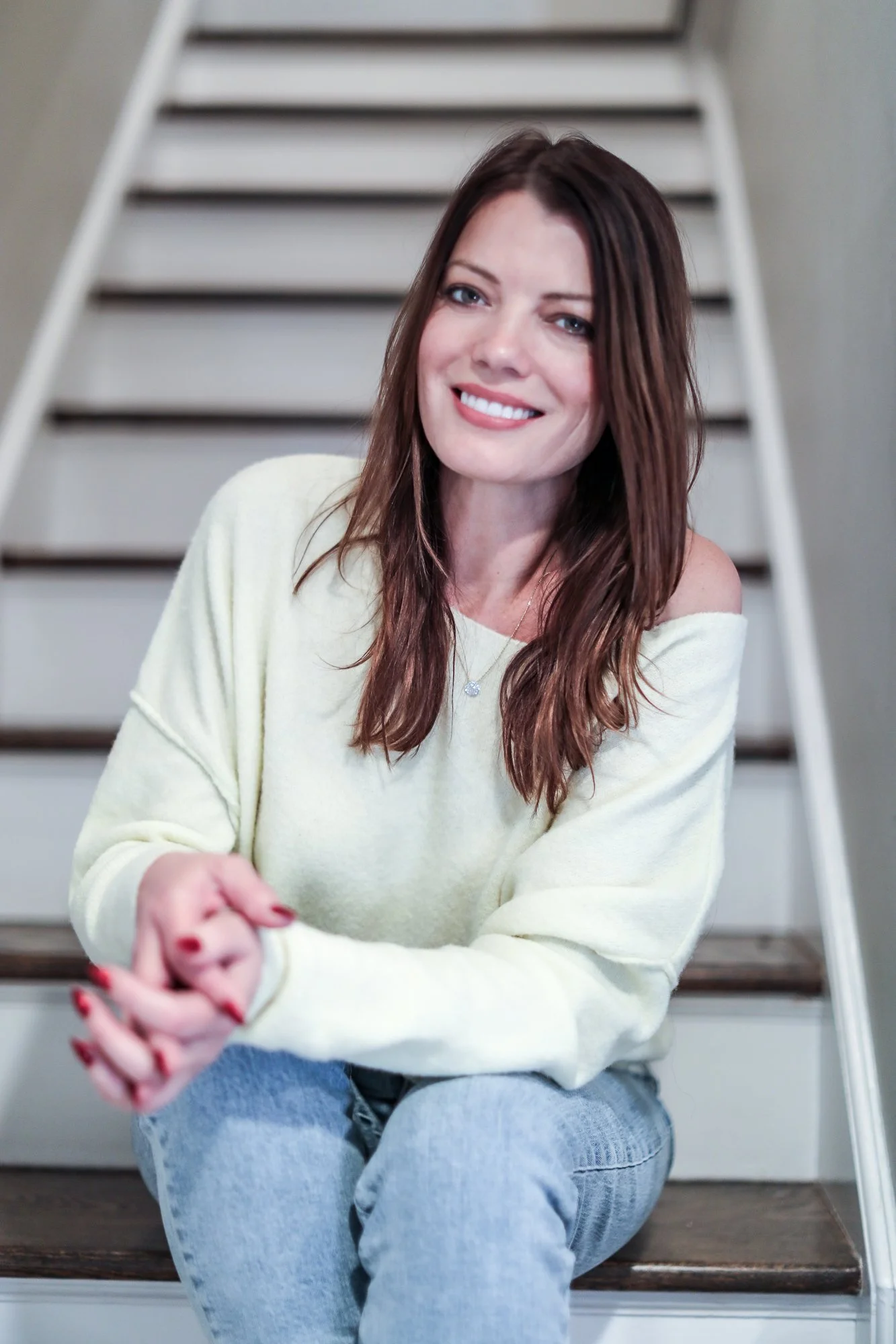 A woman with long brown hair sitting on stairs, wearing a light yellow sweater and blue jeans, smiling at the camera.