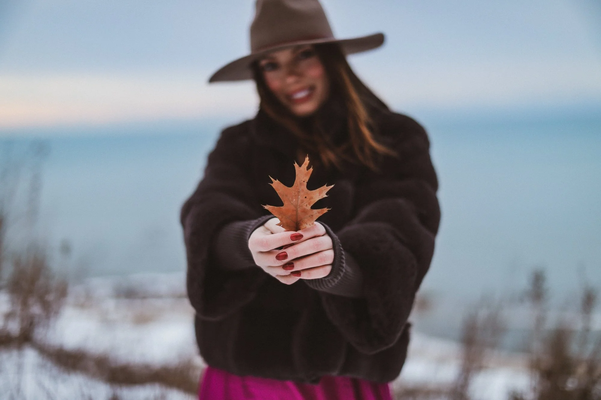 A woman wearing a brown hat and black coat holding a brown maple leaf toward the camera. She has red nail polish and is smiling, standing outdoors with a snowy landscape and a blurred body of water or sky in the background.
