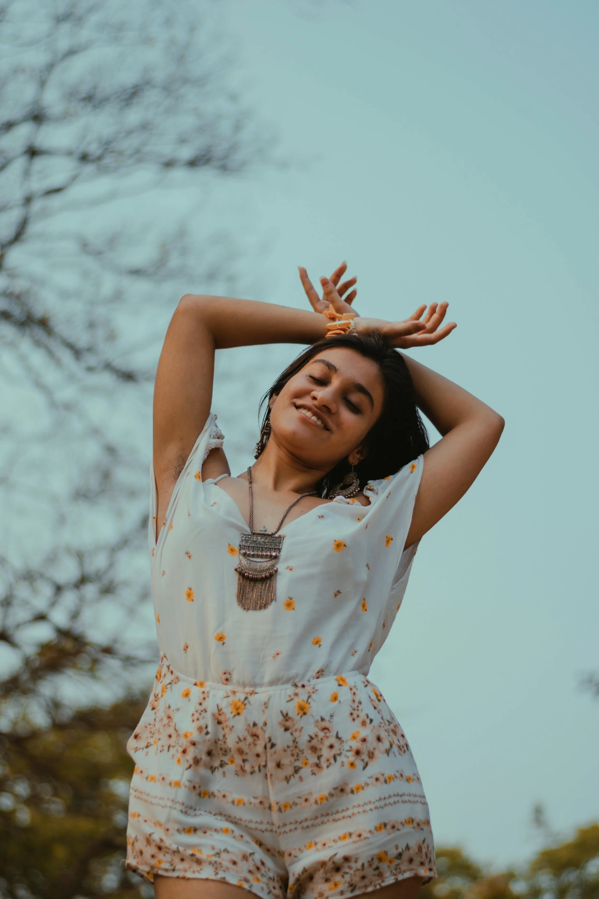 A woman in a floral romper smiling with her arms raised behind her head outdoors with trees in the background.