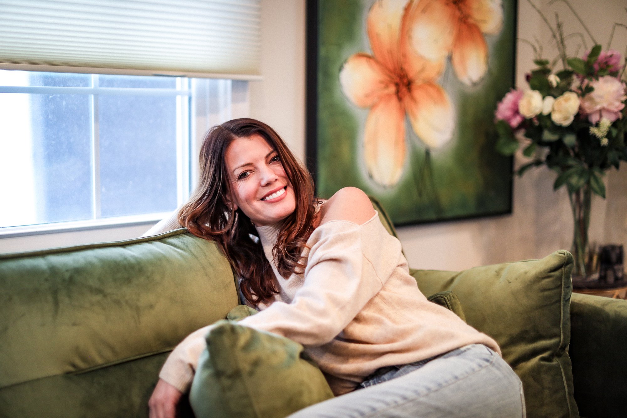 A woman with brown hair smiling and relaxing on a green sofa in a living room with a window, painting, and flowers.