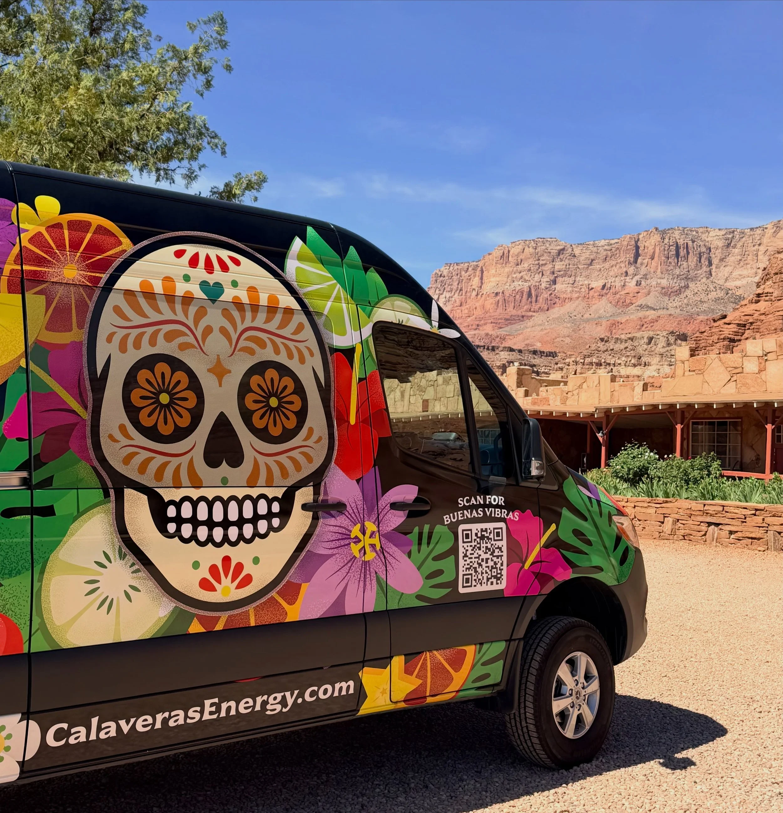 A van with colorful Day of the Dead skull and flower artwork parked against a backdrop of mountains and trees.