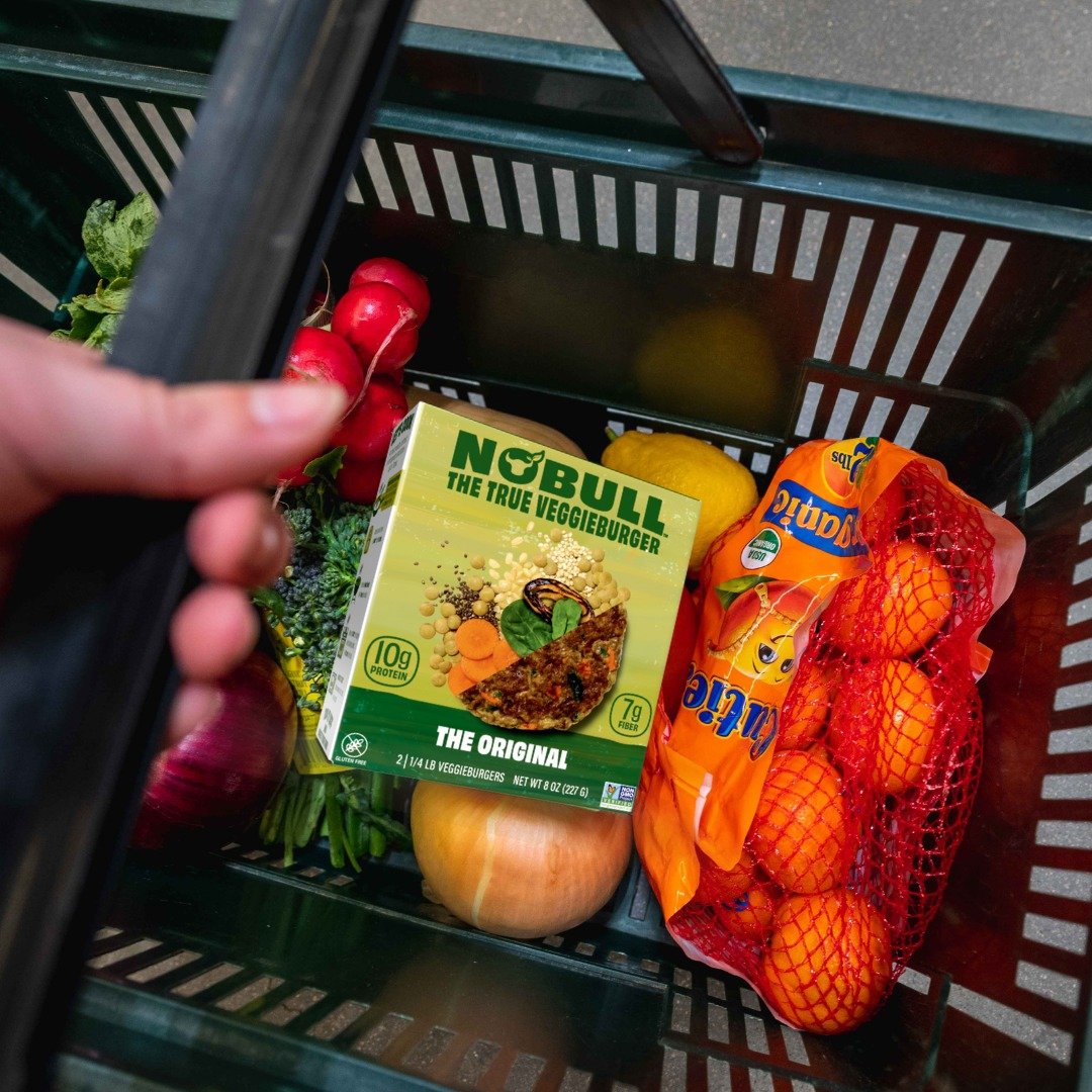 A grocery shopping cart containing a box of Nobull veggie burger, a bunch of radishes, a yellow lemon, a red apple, an orange bag of oranges, and a yellow onion.