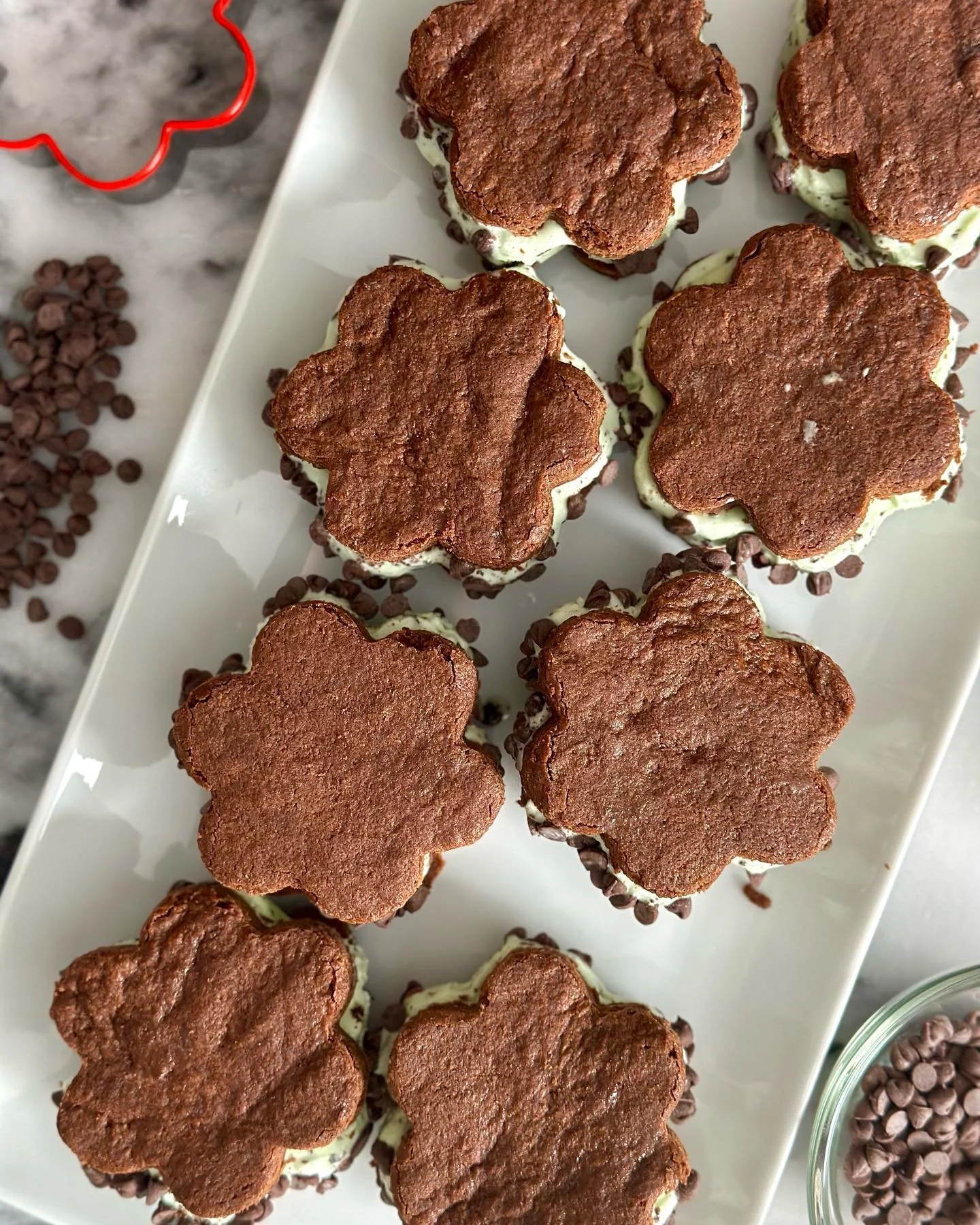 A white rectangular platter with 12 heart-shaped and flower-shaped chocolate and vanilla cream desserts topped with chocolate cookies and sprinkles, surrounded by containers of chocolate chips on a marble surface.