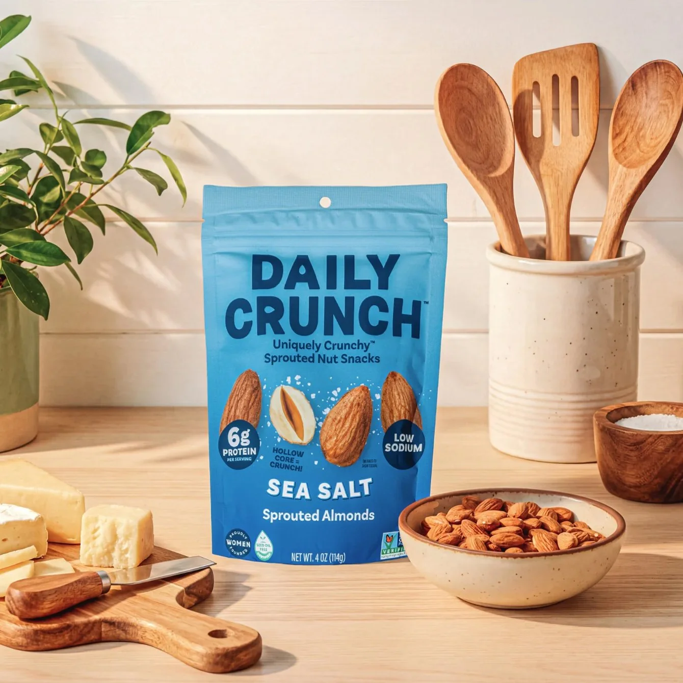 A blue bag of Daily Crunch Sea Salt Sprouted Almonds on a wooden countertop, surrounded by cheese blocks, a bowl of almonds, a wooden spoon, and a knife, with a potted plant and wooden utensils in the background.