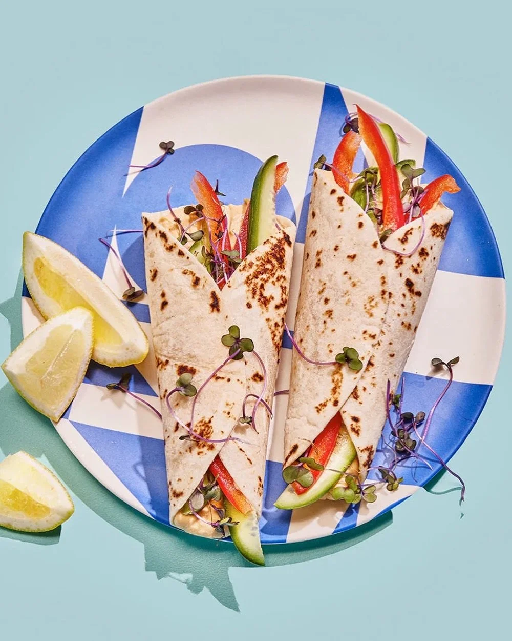 Two grilled chicken wraps with sliced vegetables inside, garnished with microgreens, served with lemon wedges on a blue and white plate.