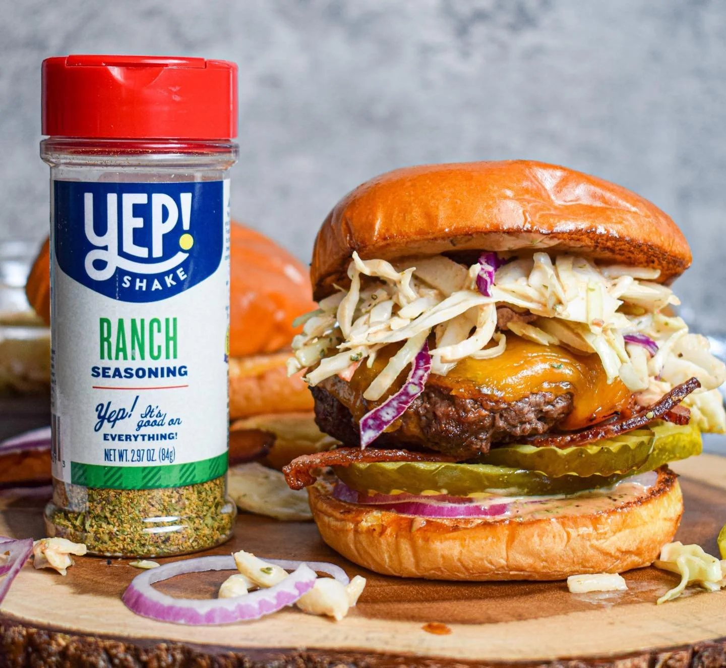 A burger with cheese, coleslaw, pickles, onions, and bacon on a bun, alongside a container of ranch seasoning.
