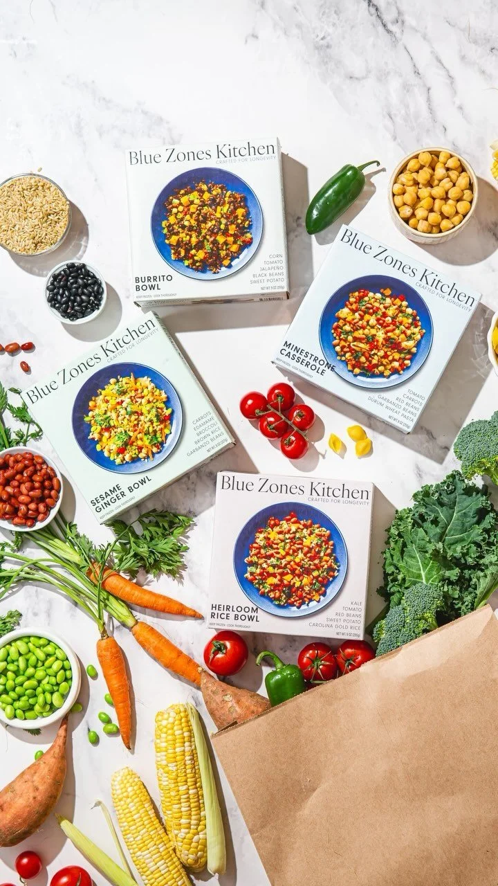 Various vegetable ingredients and Blue Zones Kitchen meal boxes on a white surface. The meal boxes include Mushroom Casserole, Sesame Ginger Bowl, Heirloom Rice Bowl, and others. Vegetables like carrots, cherry tomatoes, corn on the cob, broccoli, sweet potatoes, and a jalapeño are visible.