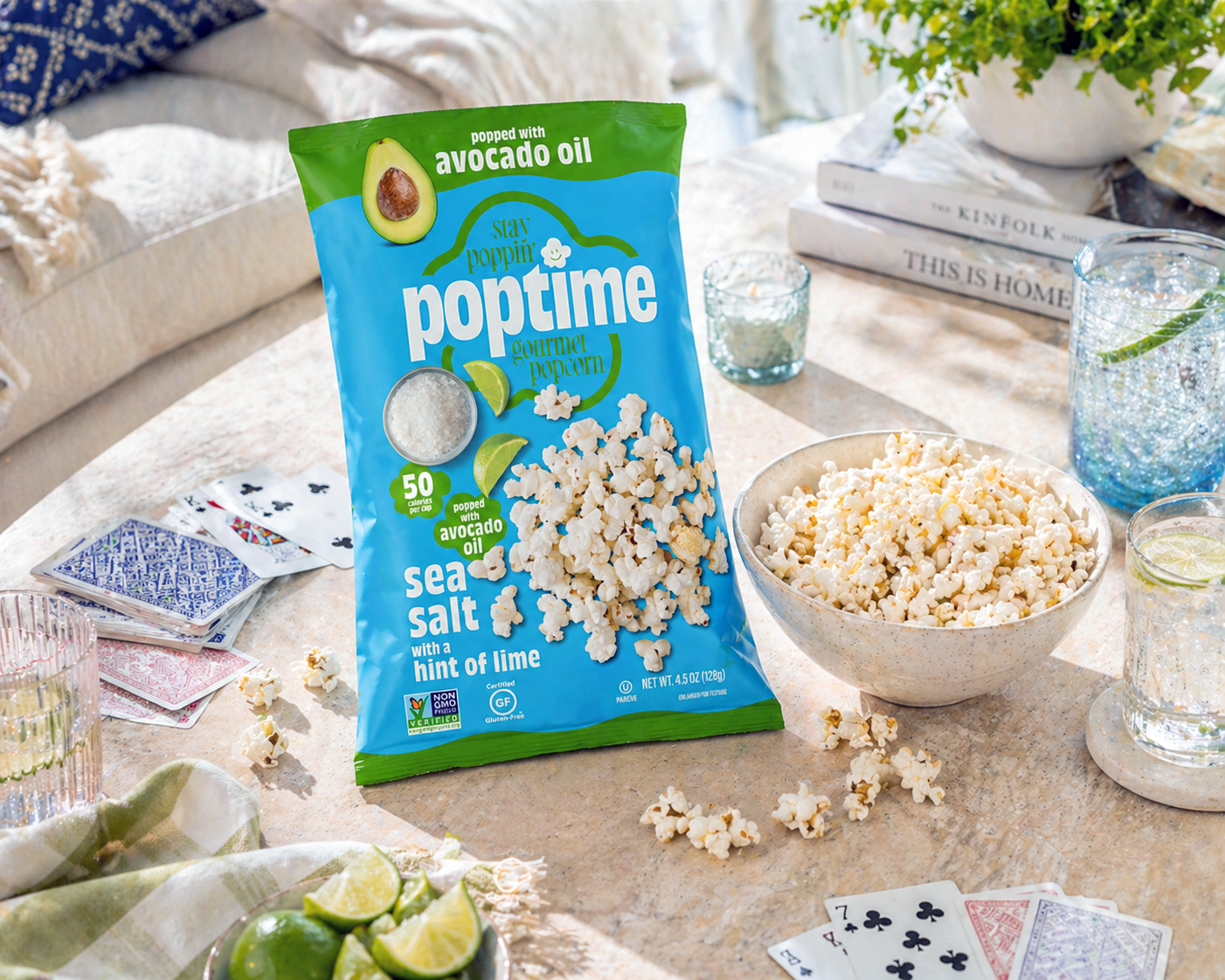 A bag of PopTime gourmet popcorn with avocado oil and sea salt on a table, surrounded by bowls of popcorn, glasses of water with lime, playing cards, and lime wedges, set for a casual gathering.