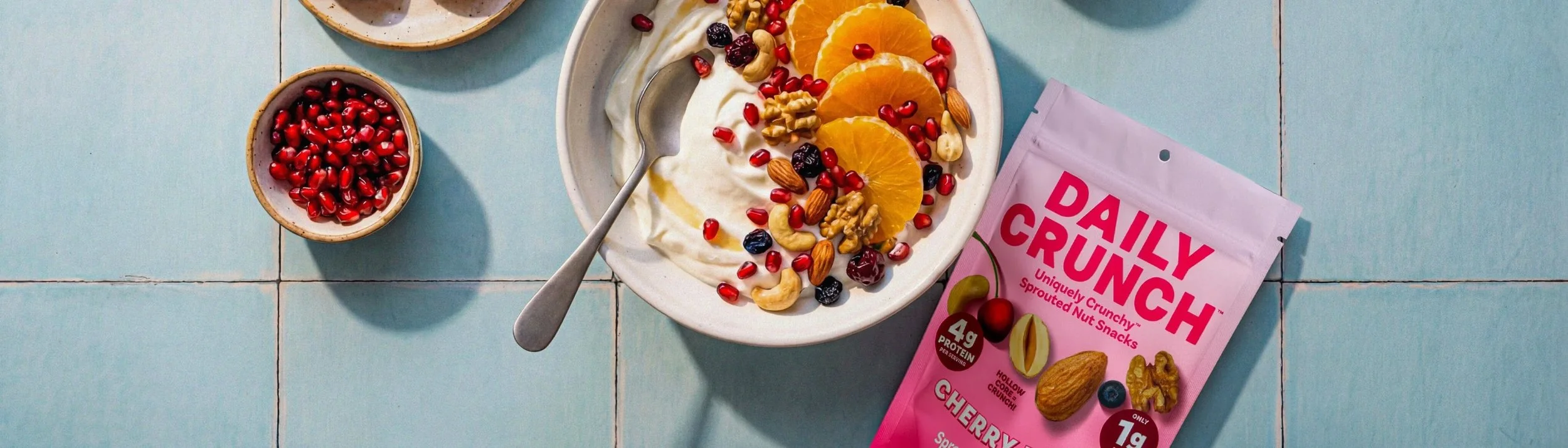 A bowl of yogurt topped with orange slices, pomegranate seeds, blueberries, cashews, almonds, and walnuts next to a pink packet of Daily Crunch cherry-flavored sprouted nut snacks on a light blue tiled surface.