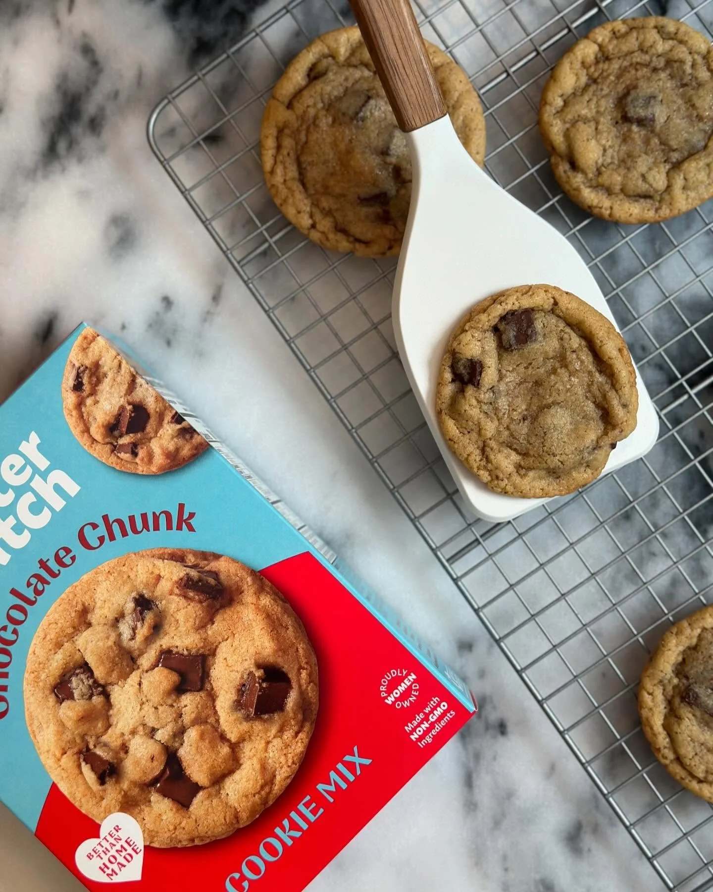 Freshly baked chocolate chip cookies on a cooling rack and a cookie mix box on a marble countertop.