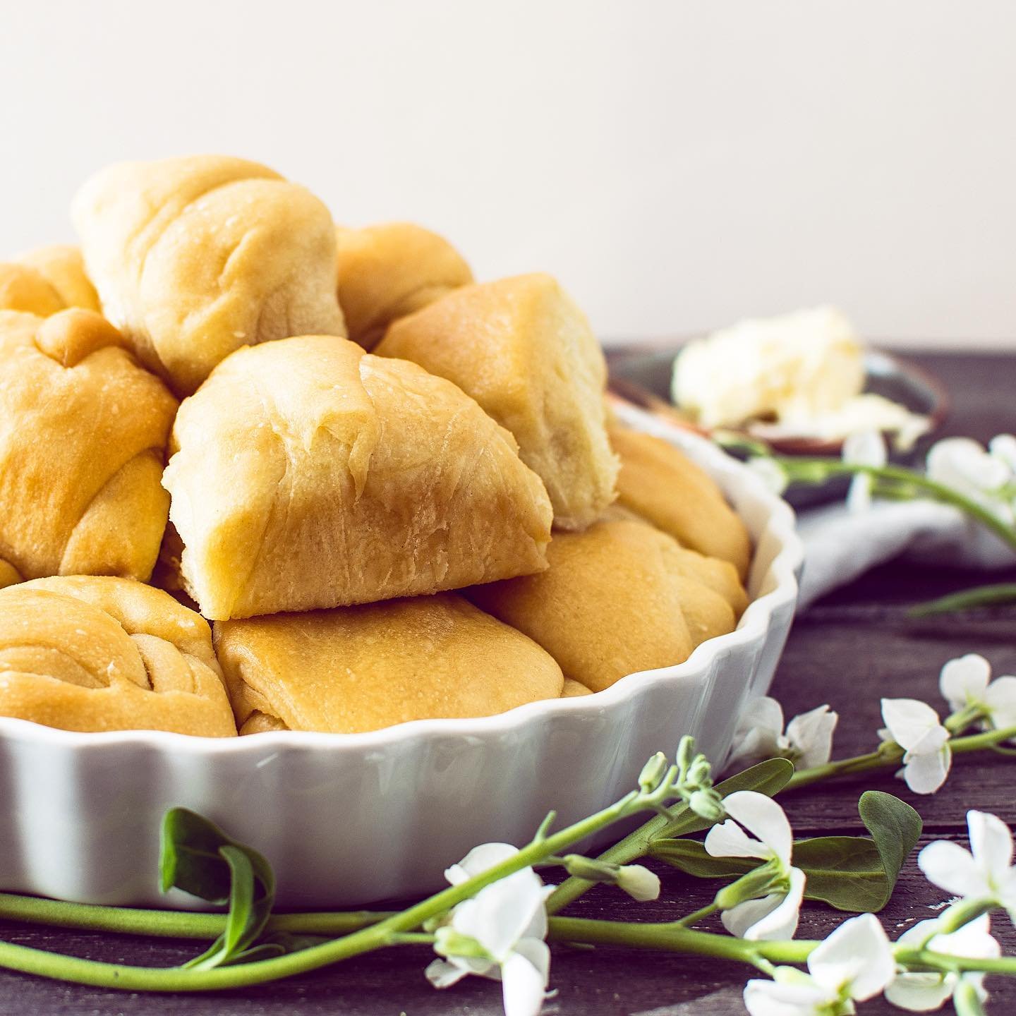A white bowl filled with golden, baked bread rolls, placed on a dark wooden surface with white flowers and green leaves around it.