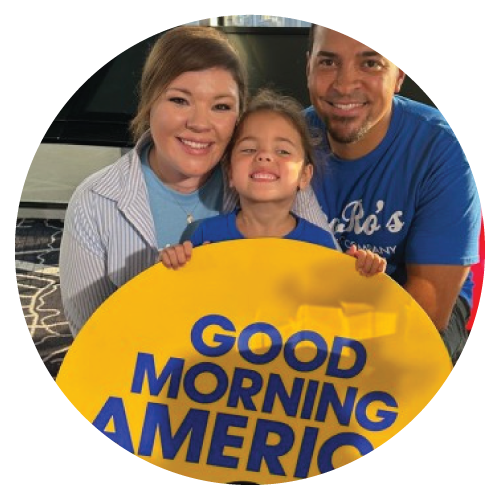 A family of three smiling and holding a yellow sign that reads "Good Morning America" in front of a black vehicle.