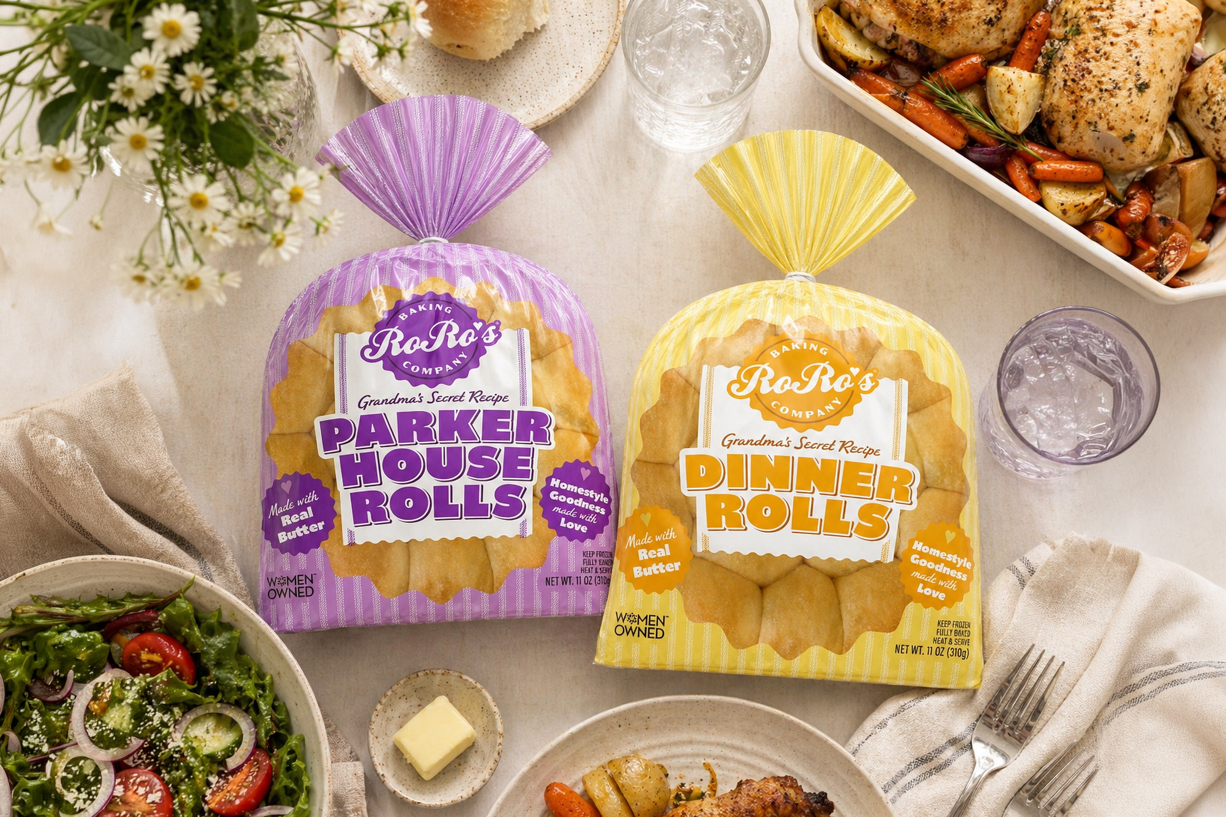 Two bags of RoRo's Dinner Rolls, one with purple packaging labeled "Parker House Rolls" and the other with yellow packaging labeled "Dinner Rolls," on a beige tablecloth surrounded by various dishes including a salad, cooked chicken with vegetables, a drinking glass with ice water, and a small block of butter.