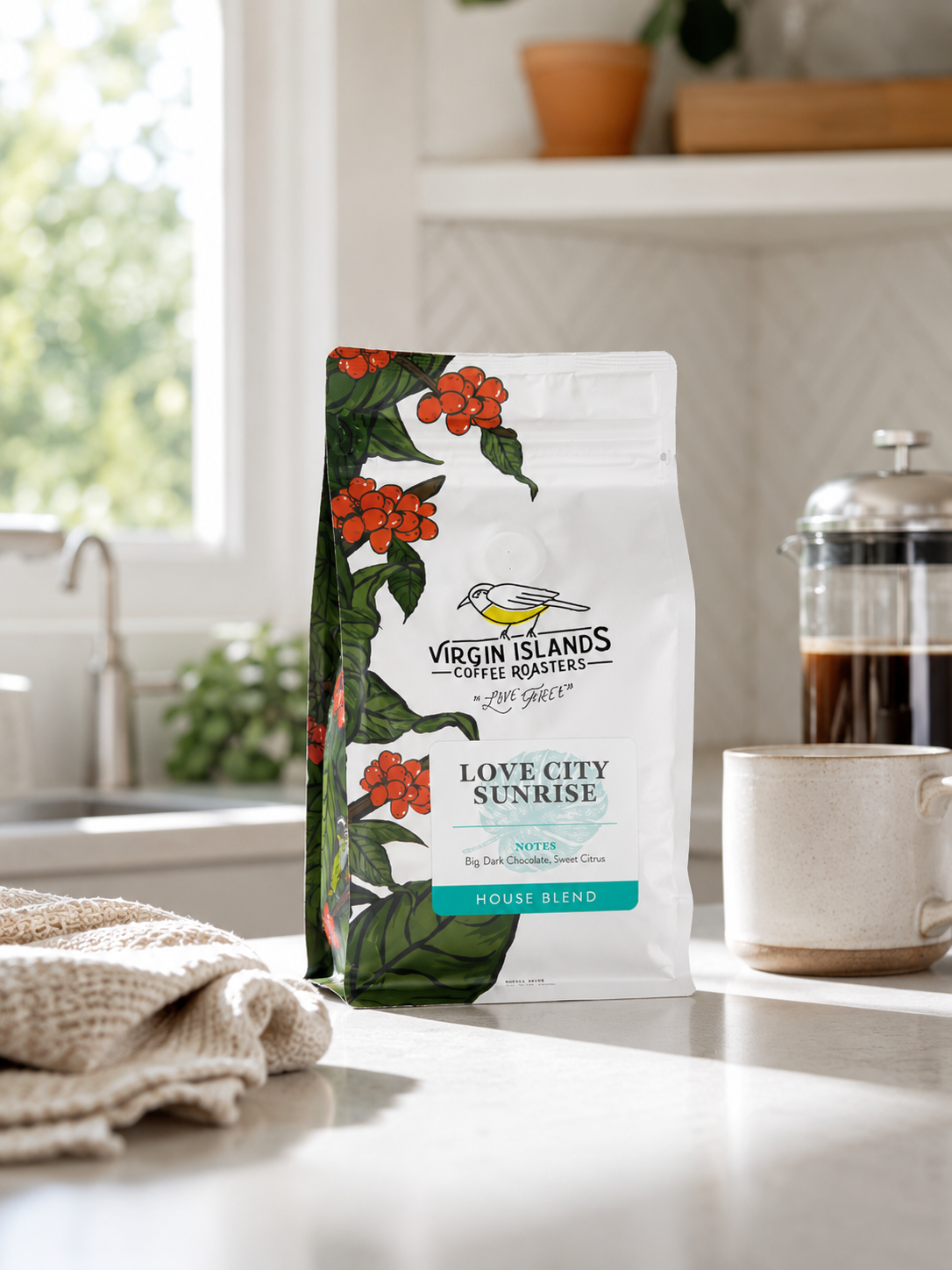 A bag of Virgin Islands Coffee Roasters Love City Sunrise house blend coffee on a kitchen counter, with a mug, French press, towel, and window in the background.